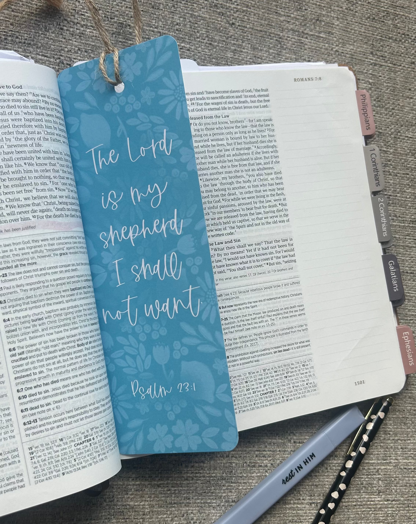 A blue patterned Bible bookmark with white script text reading 'The Lord is my shepherd I shall not want Psalm 78:1' resting on an open Bible. The bookmark is styled in a flat-lay with a gray pen and a black-and-white polka dot pen on a textured gray fabric background.