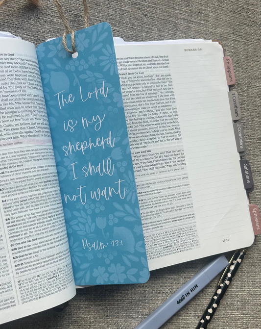 A blue patterned Bible bookmark with white script text reading 'The Lord is my shepherd I shall not want Psalm 78:1' resting on an open Bible. The bookmark is styled in a flat-lay with a gray pen and a black-and-white polka dot pen on a textured gray fabric background.