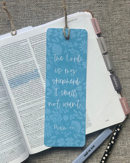 A blue patterned bookmark with white script text reading 'The Lord is my shepherd I shall not want Psalm 23:1' hanging from a book, styled on a gray textured surface with an open Bible, pen, and pencil.
