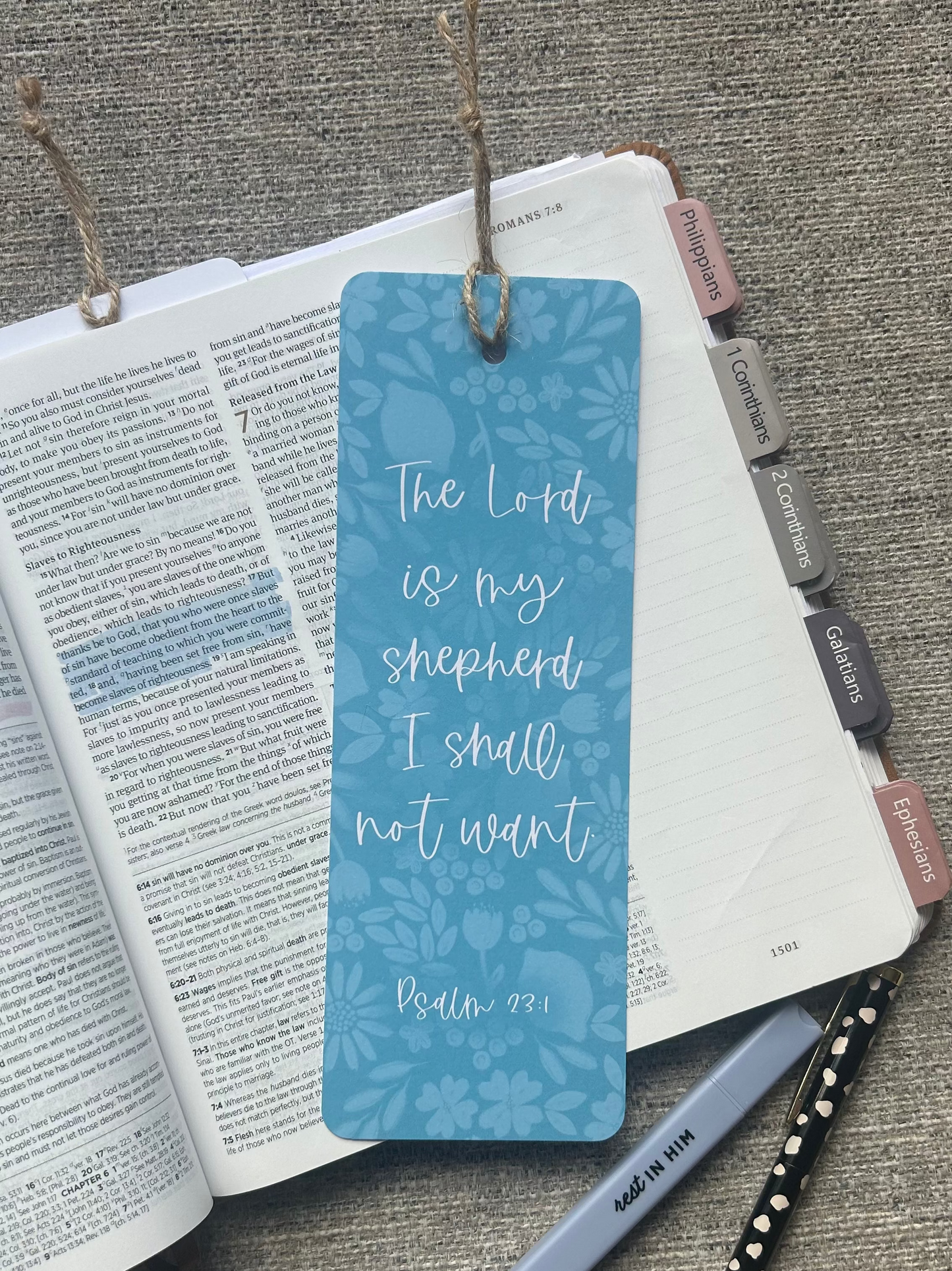 A blue patterned bookmark with white script text reading 'The Lord is my shepherd I shall not want Psalm 23:1' hanging from a book, styled on a gray textured surface with an open Bible, pen, and pencil.