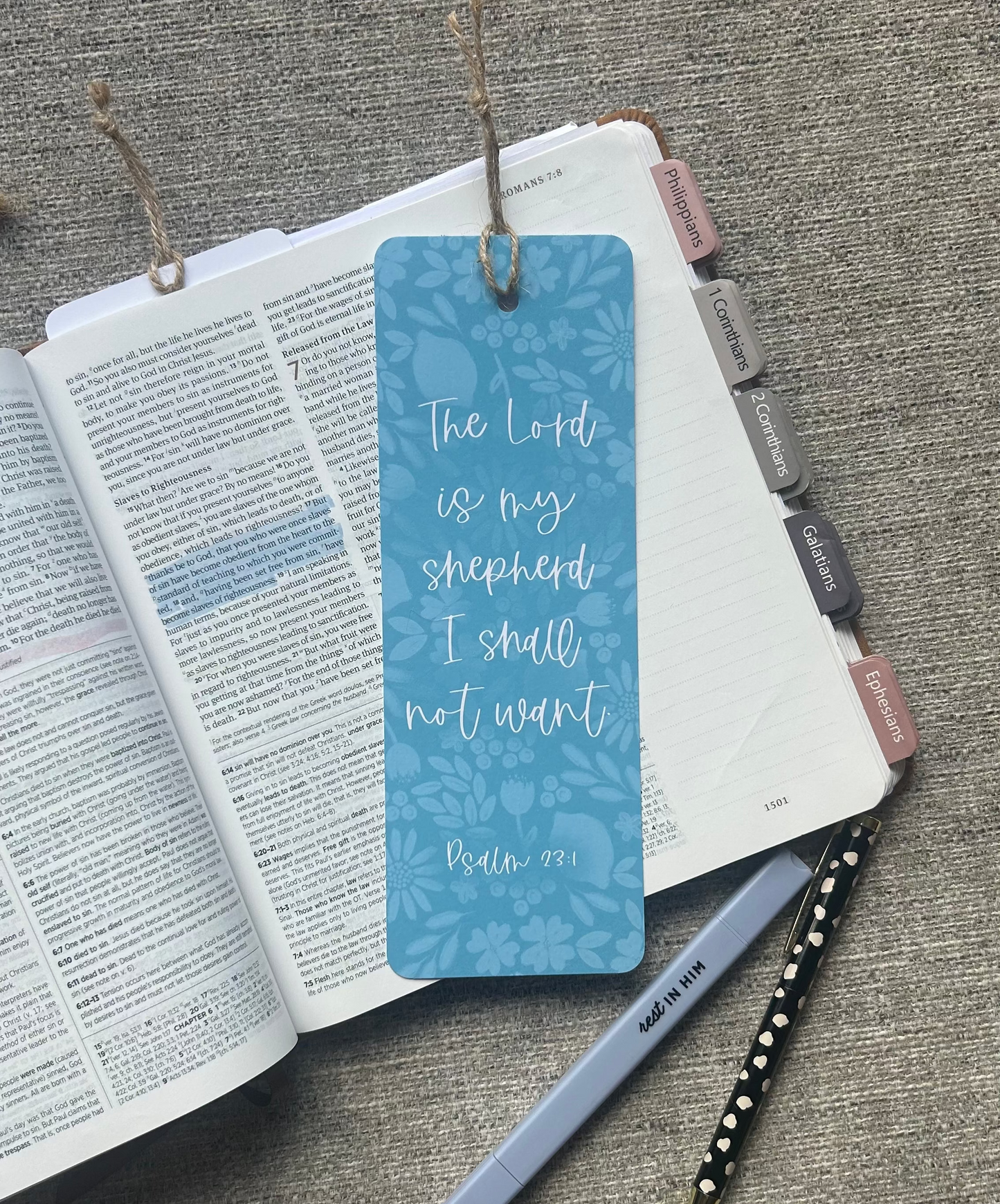 A blue rectangular bookmark with white script text reading 'The Lord is my shepherd I shall not want Matt 18:14' hangs from a twine string over an open book, with pens nearby on a textured gray fabric background.