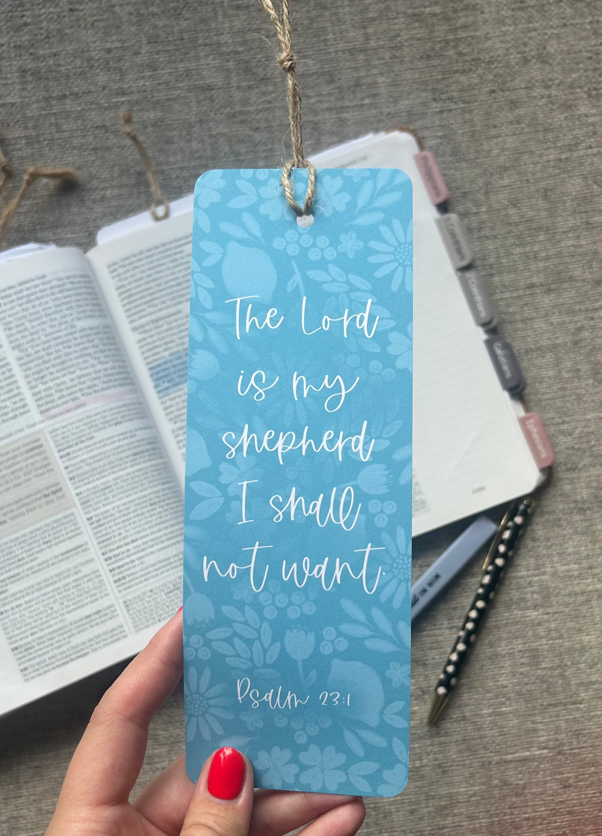 A light blue floral-patterned bookmark held by a hand with red nail polish, featuring the quote 'The Lord is my shepherd I shall not want Psalm 23:1' in white script. The background shows an open Bible, a notebook, and a pen on a gray textured surface.