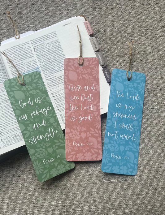 Three decorative bookmark tags with Bible verse quotes are arranged on top of an open book against a gray textured fabric background. The tags are green, dusty pink, and blue, each with a subtle patterned background and white script text.