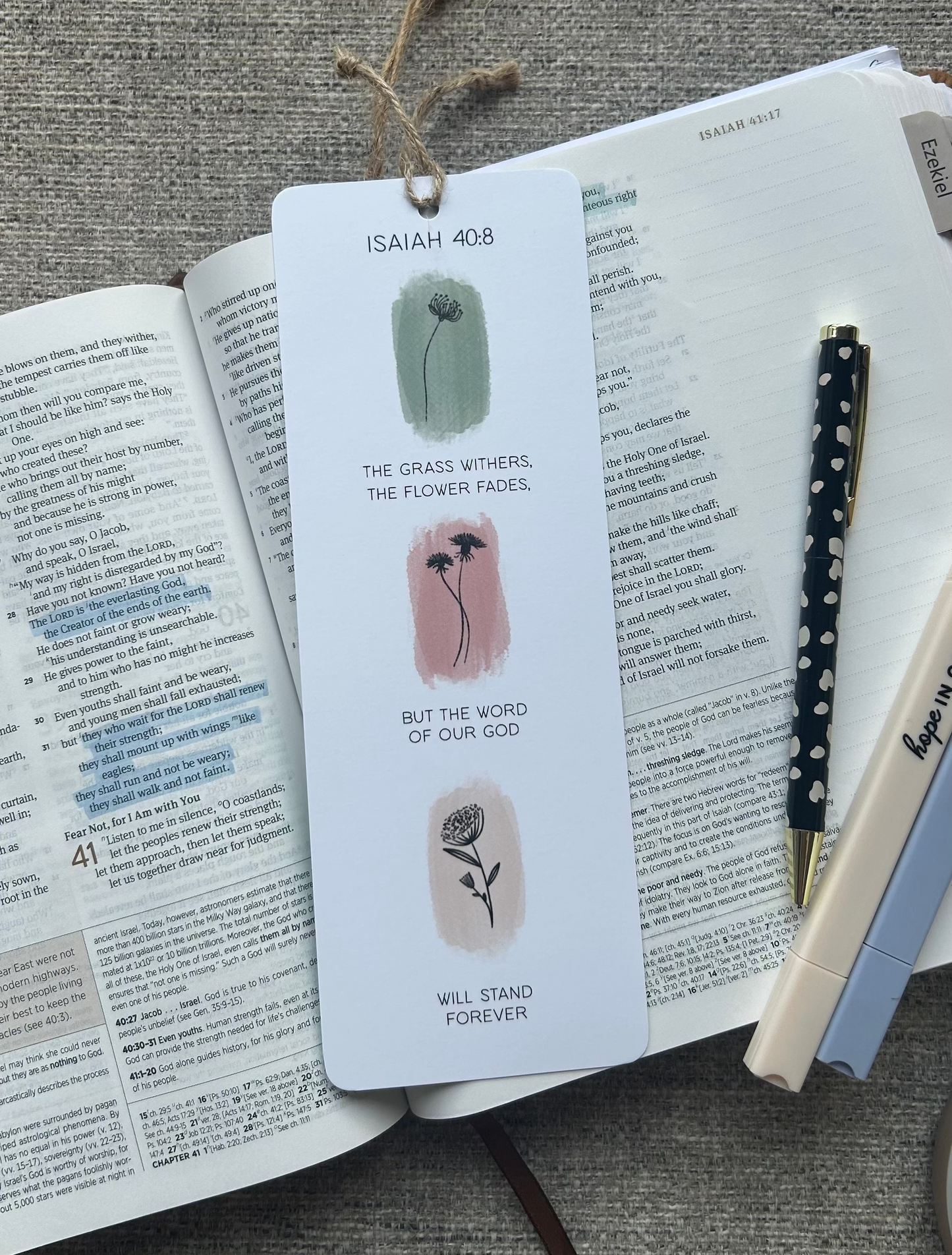 A white paper bookmark with illustrations and text from Isaiah 40:8 lies on an open book, styled with a pen and another pen on a textured gray fabric background.
