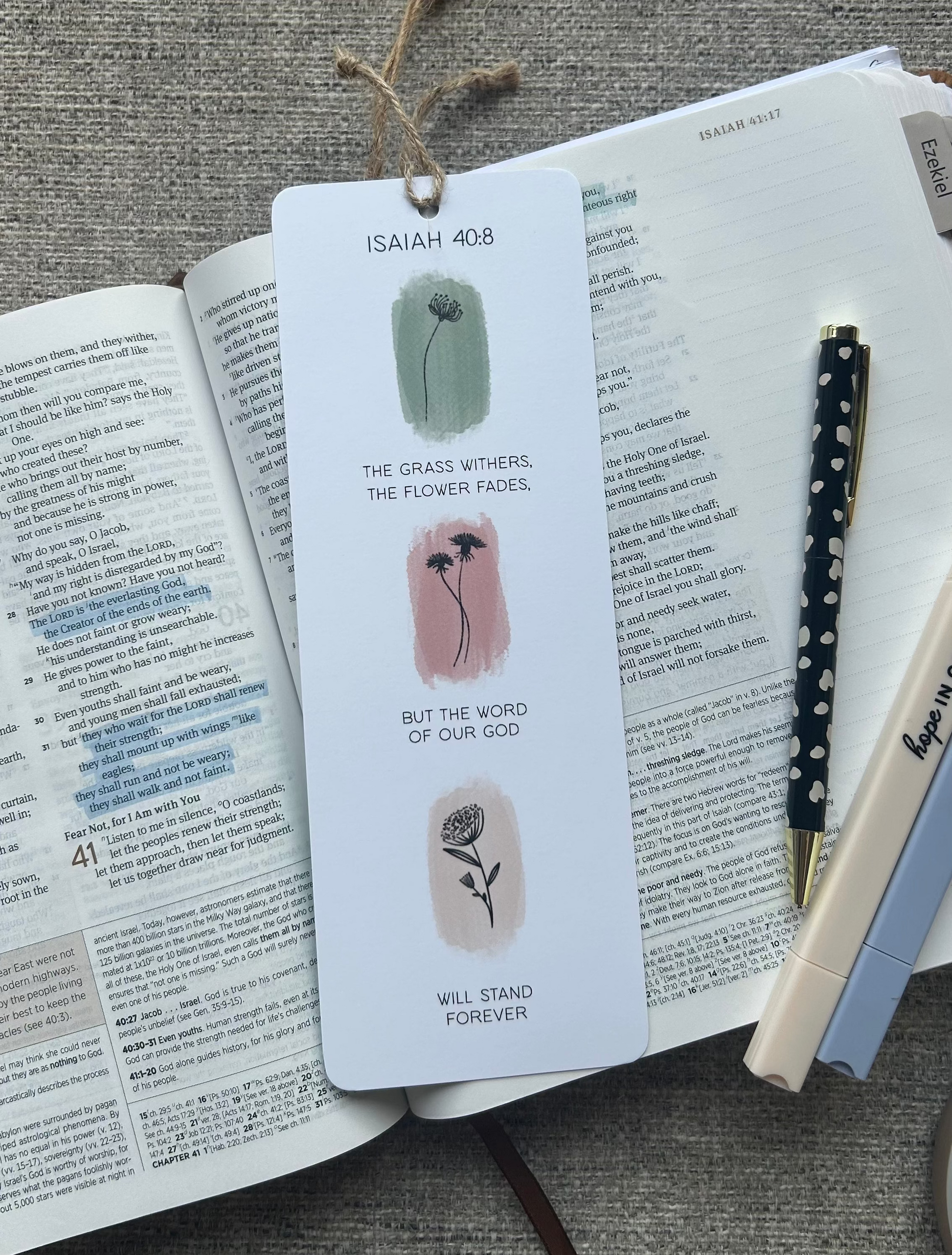 A white paper bookmark with illustrations and text from Isaiah 40:8 lies on an open book, styled with a pen and another pen on a textured gray fabric background.