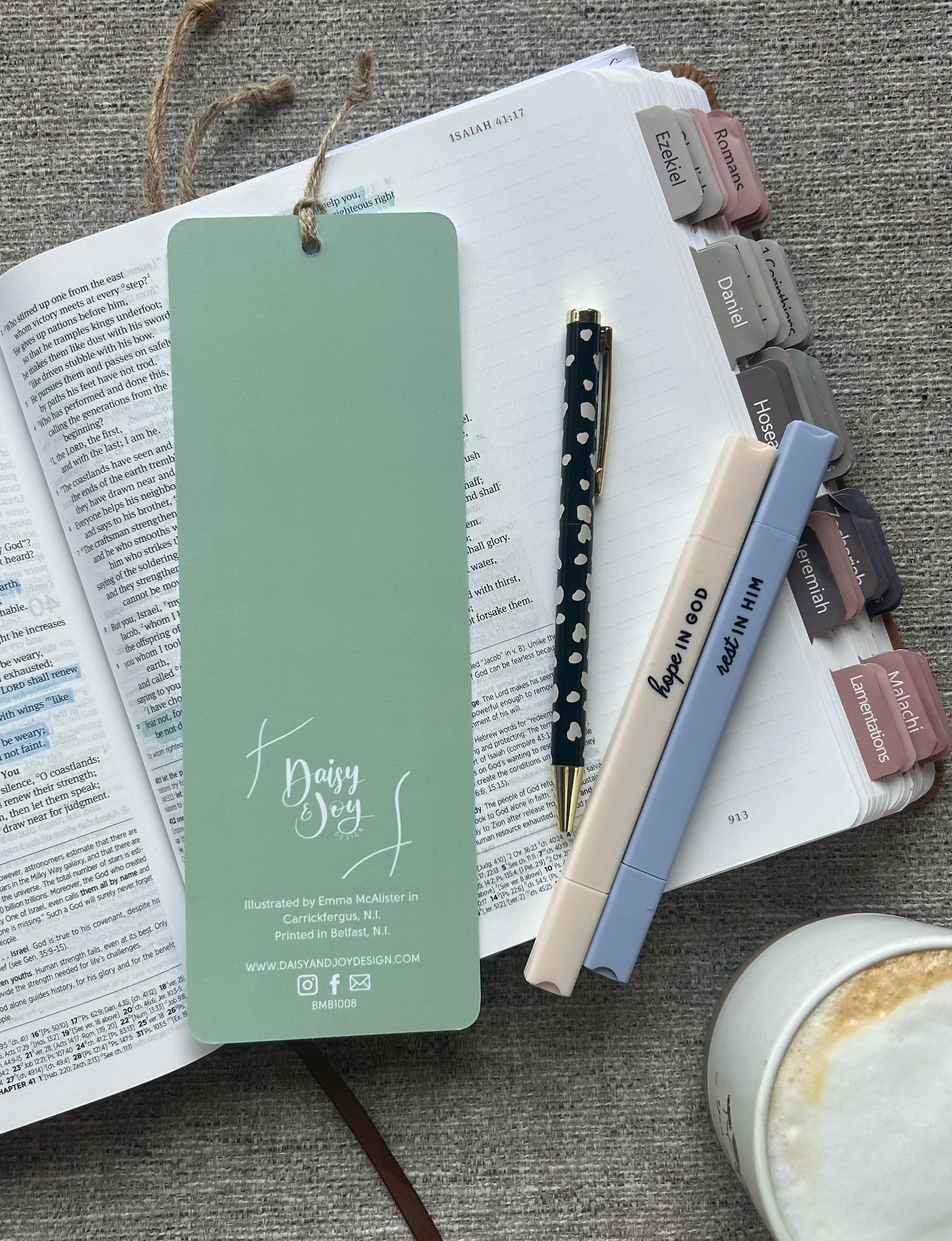 A sage green paper bookmark with a hanging string and 'Daily Joy' script, styled on an open book with pens and a coffee cup on a gray textured surface.