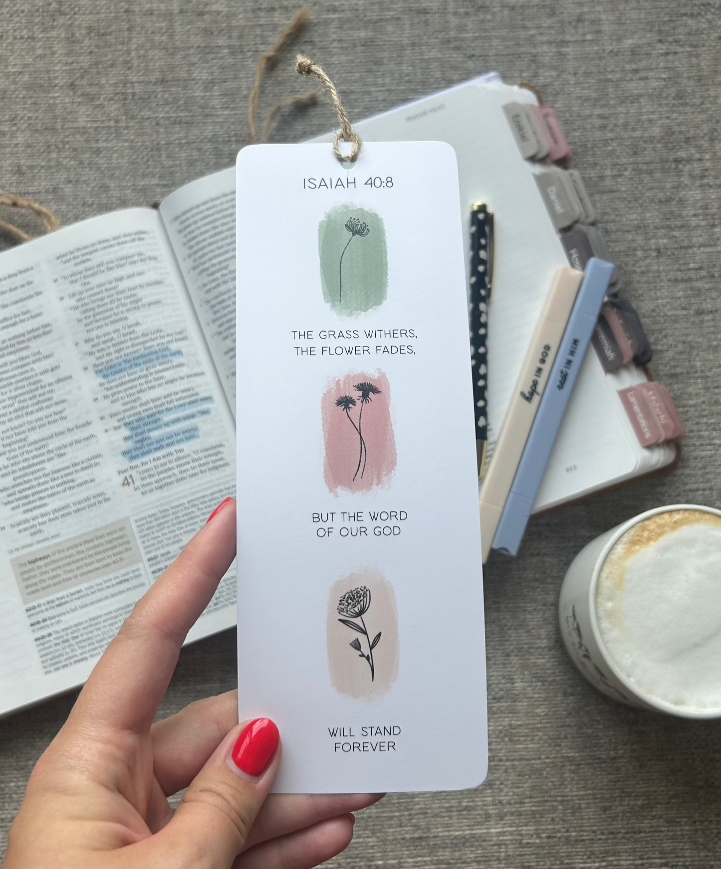 A hand with red nail polish holds a white rectangular bookmark card featuring illustrations and text from Isaiah 40:1-8. The background shows a styled desk setup with an open book, a notebook, pens, and a candle on a gray fabric surface.