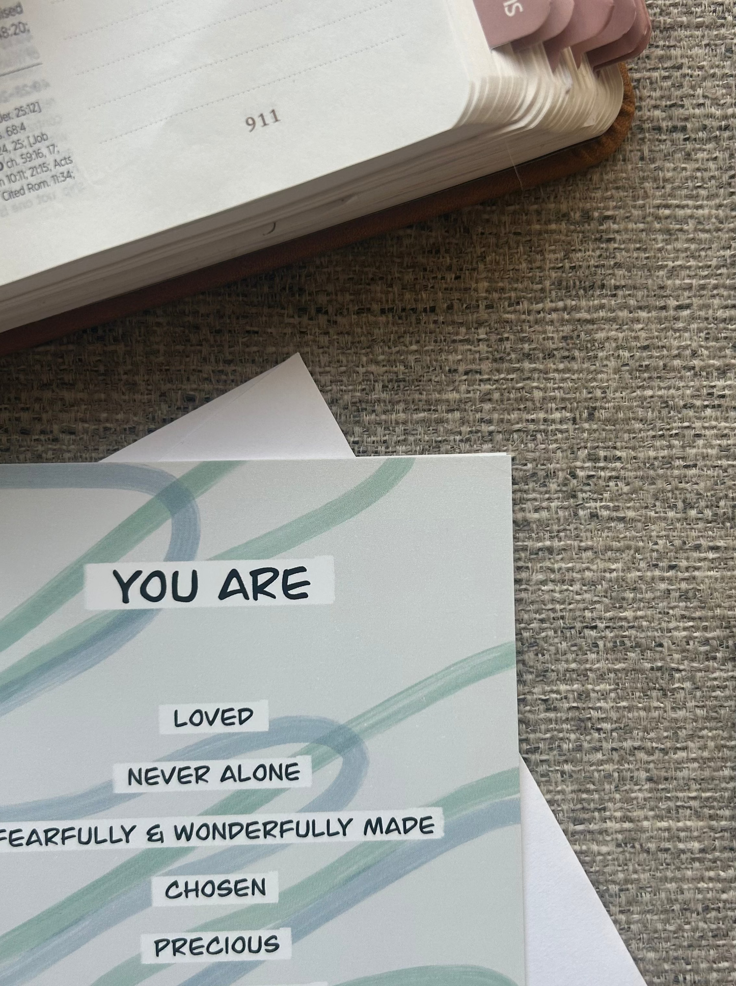 A printed affirmation card with the text 'YOU ARE' and supporting phrases like 'LOVED', 'NEVER ALONE', and 'CREATED FOR A PURPOSE' placed on top of a Bible, photographed on a textured fabric surface.