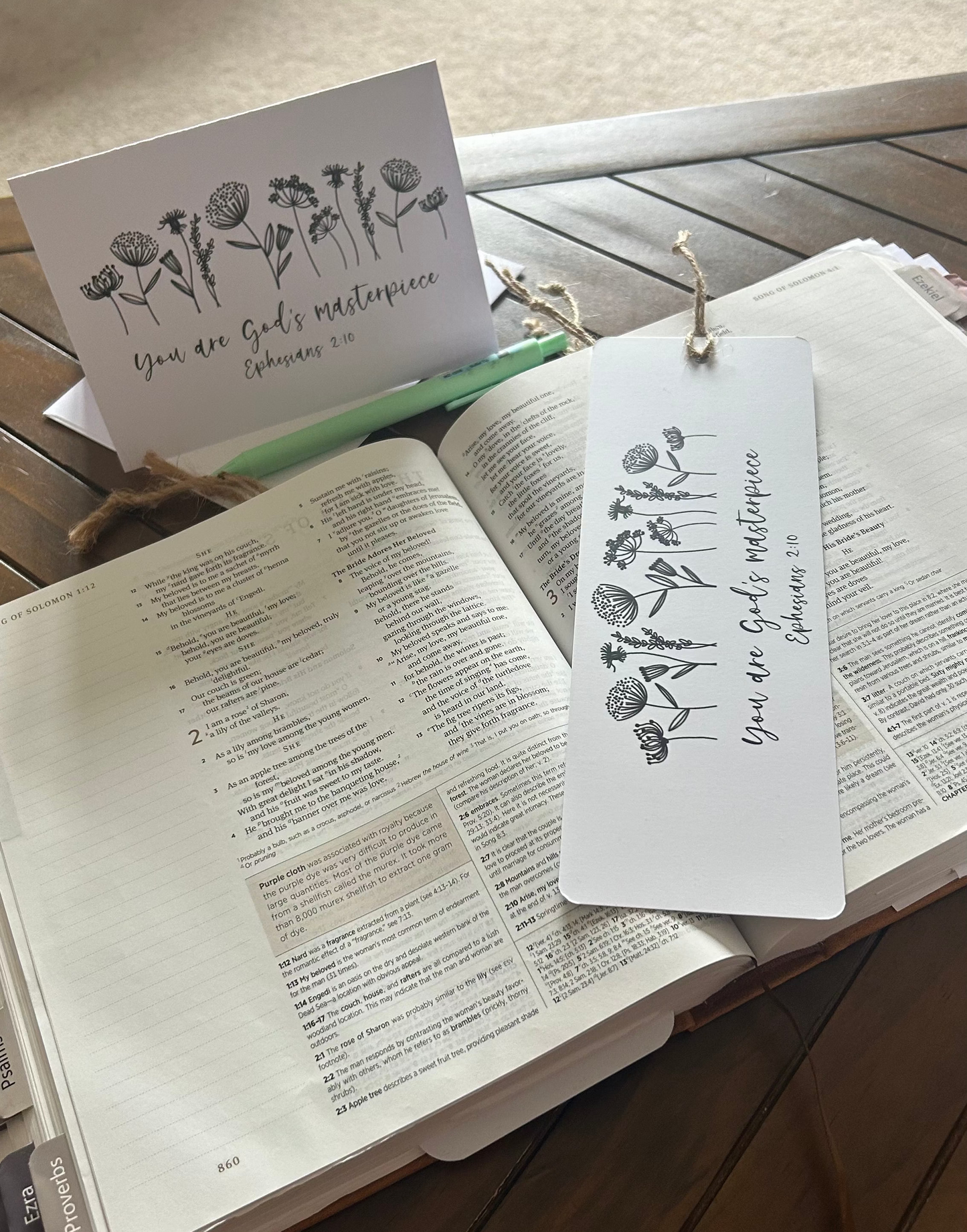 A white bookmark card with black floral line art and the words 'You are God's masterpiece' rests on an open Bible on a wooden table, next to a matching card and a green pen.