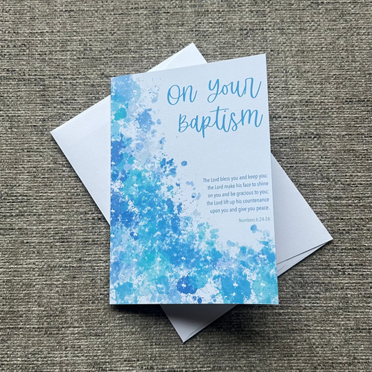 A baptism announcement card with a blue watercolor splatter design and white envelope, displayed on a textured gray fabric background.