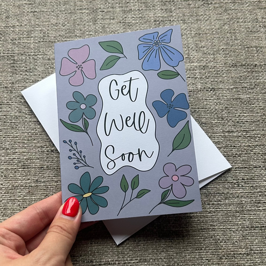 A hand with red nail polish holds a square 'Get Well Soon' greeting card featuring a lavender background and colorful floral illustrations, with a white envelope behind it on a textured gray fabric surface.