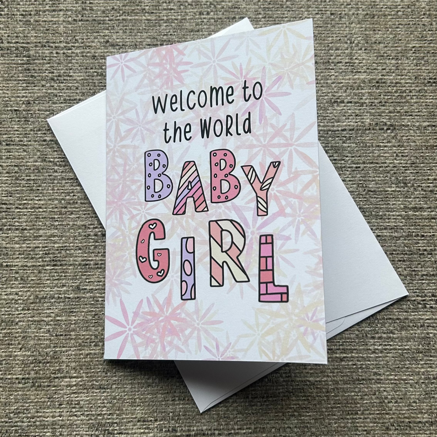 A square baby shower card reading 'Welcome to the World BABY GIRL' in colorful patterned letters, placed on top of a white envelope on a textured beige fabric background.