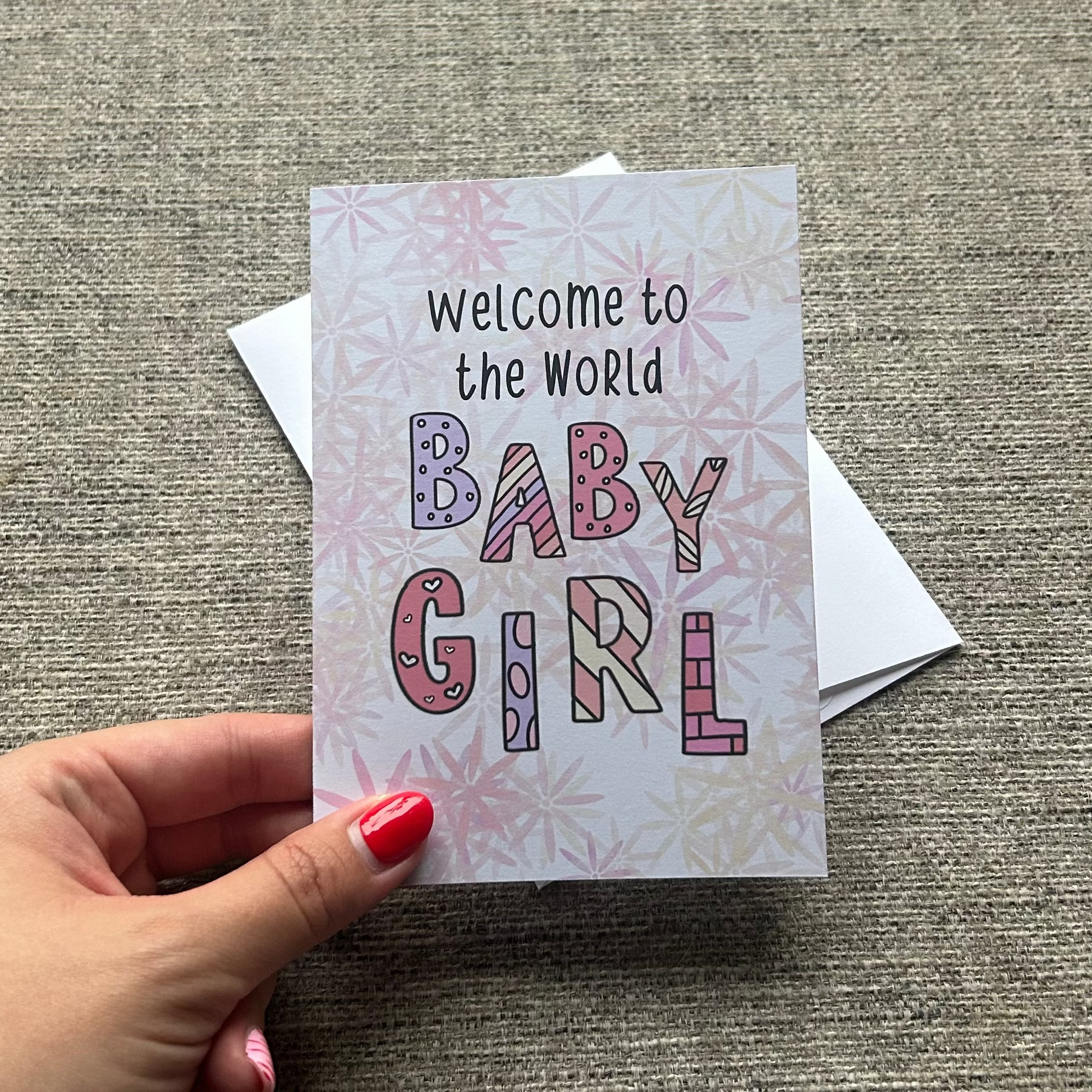 A hand with pink nail polish holds a square baby shower card that reads 'Welcome to the World BABY GIRL' in colorful patterned letters, with a white envelope behind it on a textured gray fabric background.