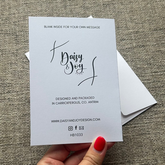 A hand with red nail polish holds a white custom business card featuring a black script logo that reads 'Dairy Soy'. Behind the card, a white envelope is visible on a textured gray fabric background.