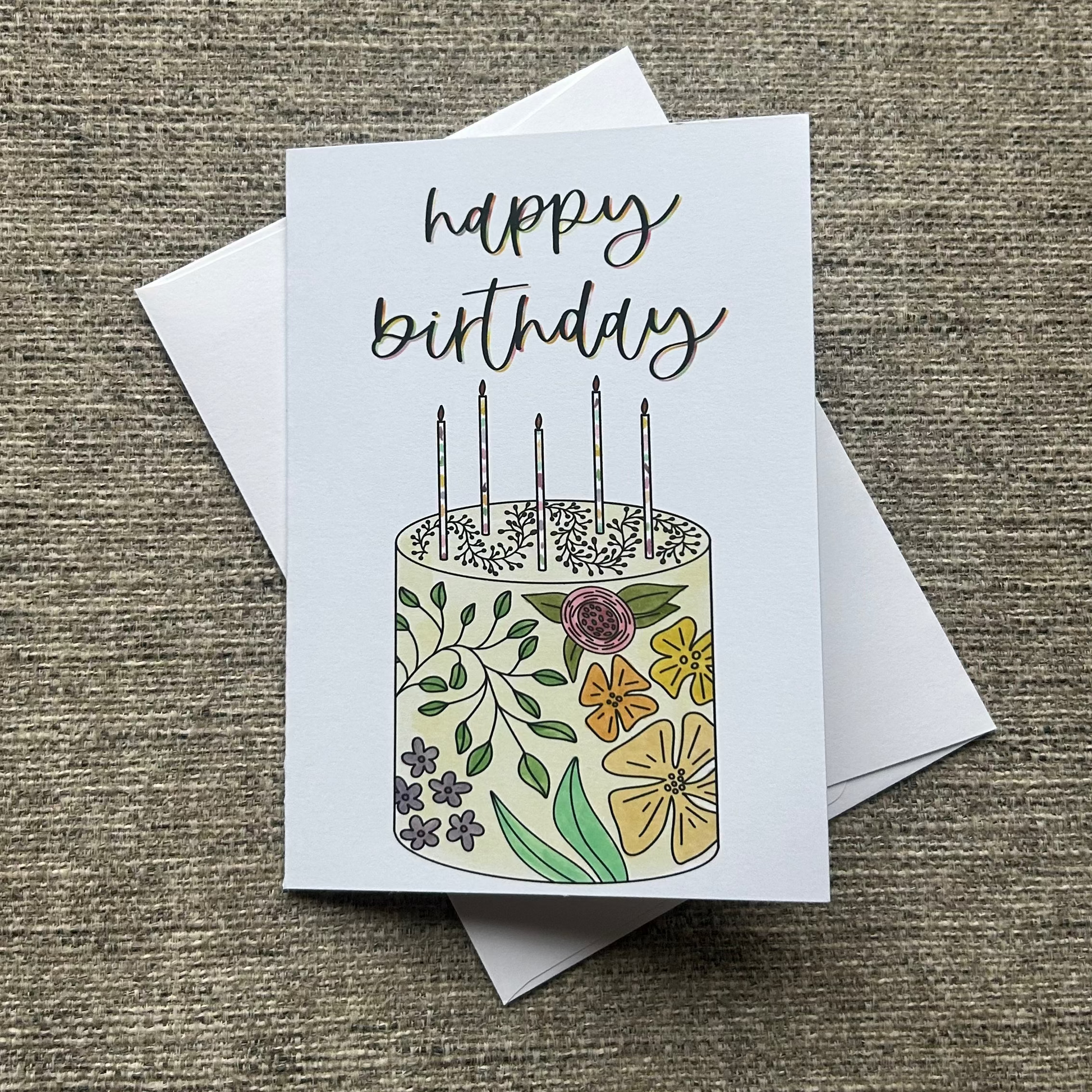 A square birthday card with a white envelope, placed on a textured beige fabric background. The card features a hand-drawn illustration of a birthday cake decorated with flowers and green leaves, topped with five candles, and the words 'happy birthday' written in black script.