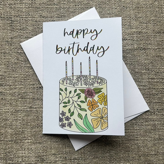 A square birthday card with a white envelope, placed on a textured beige fabric background. The card features a hand-drawn illustration of a birthday cake decorated with flowers and green leaves, topped with five candles, and the words 'happy birthday' written in black script.