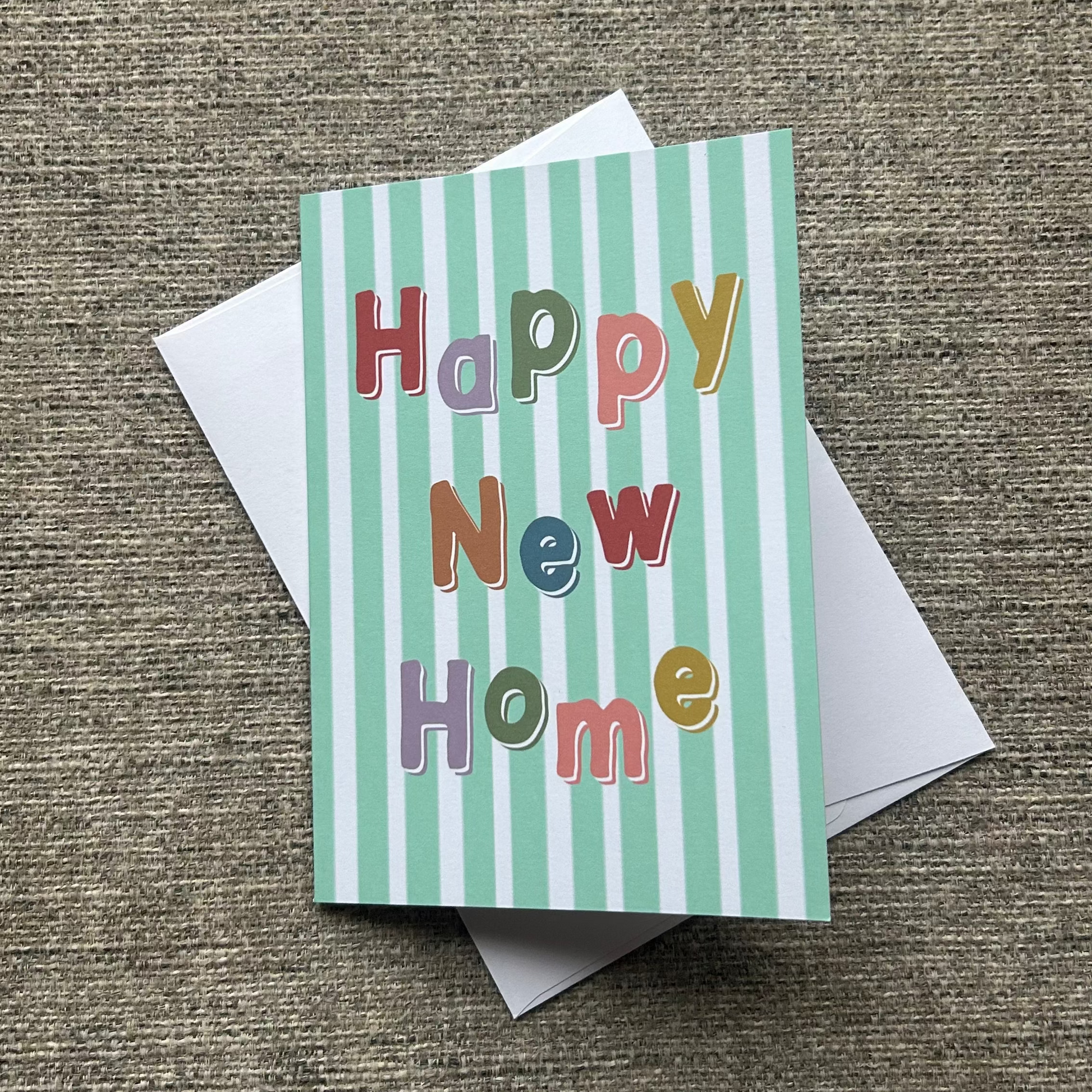 A square greeting card with pastel green and white vertical stripes and the words 'Happy New Home' in colorful raised letters, placed on top of a white envelope over a textured beige fabric background.