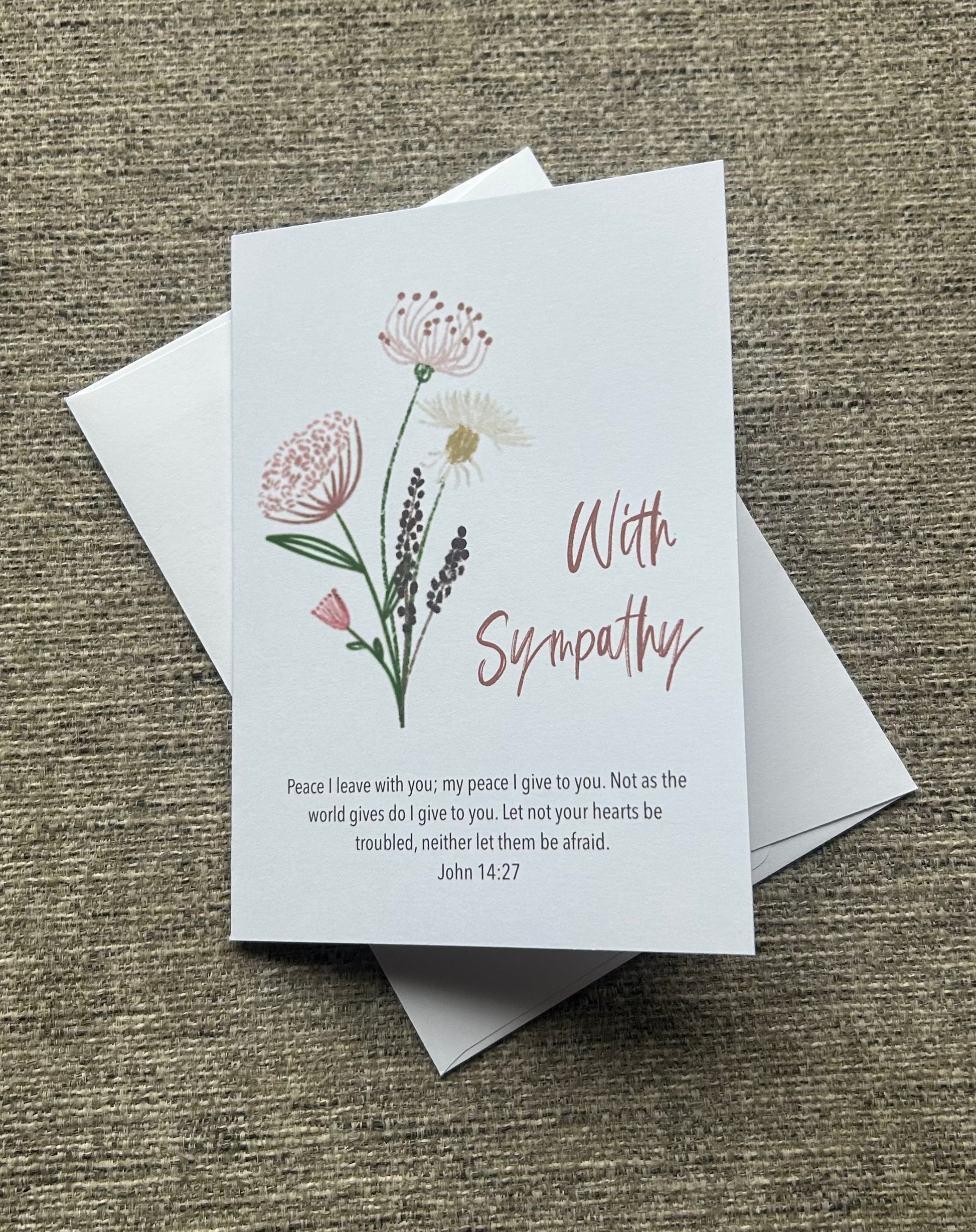 A sympathy greeting card with a white background, featuring delicate pink and green floral illustrations, the words 'With Sympathy' in pink script, and a short Bible verse printed below. The card is displayed on top of a white envelope on a textured brown fabric background.