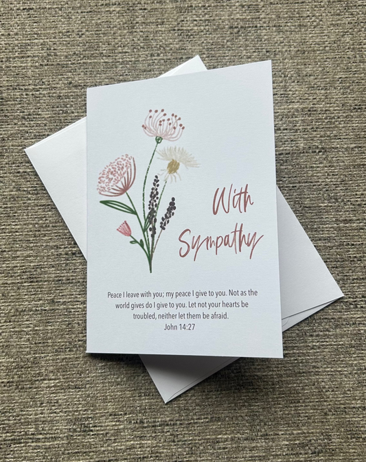 A sympathy greeting card with a white background, featuring delicate pink and green floral illustrations, the words 'With Sympathy' in pink script, and a short Bible verse printed below. The card is displayed on top of a white envelope on a textured brown fabric background.