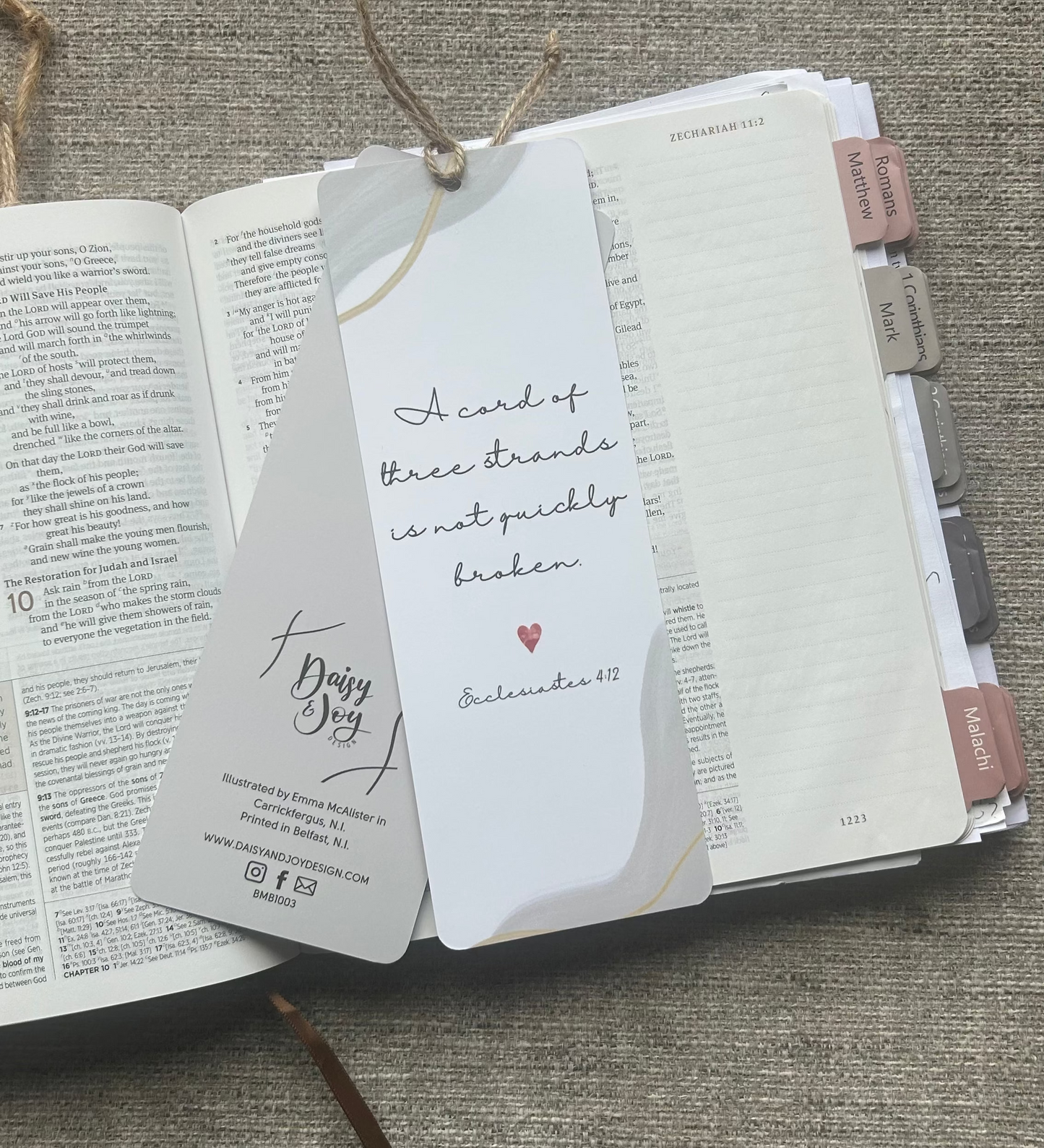 A white paper bookmark card with handwritten text and a small red heart, attached to a Bible bookmark ribbon, laid over an open book on a textured gray fabric background.