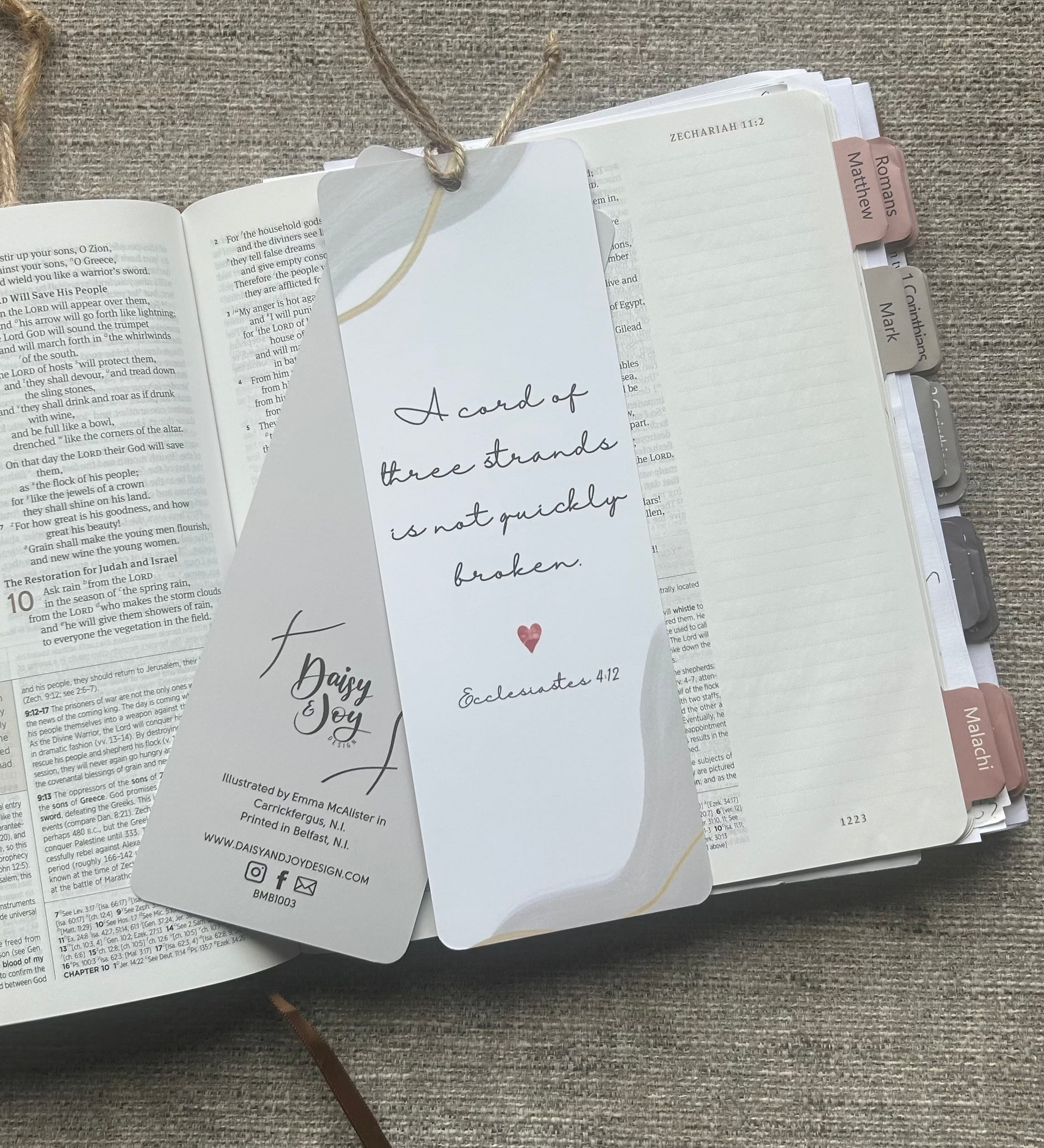 A white paper bookmark card with handwritten text and a small red heart, attached to a Bible bookmark ribbon, laid over an open book on a textured gray fabric background.