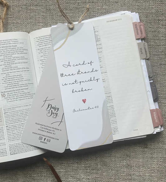 A white paper bookmark card with handwritten text and a small red heart, attached to a Bible bookmark ribbon, laid over an open book on a textured gray fabric background.