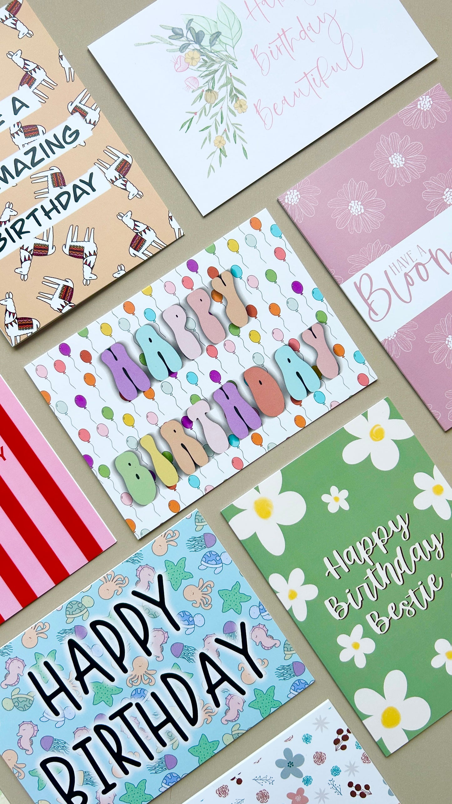 A flat-lay arrangement of multiple colorful birthday greeting cards on a neutral beige background, featuring various designs including floral illustrations, polka dots, daisies, and cartoon characters.