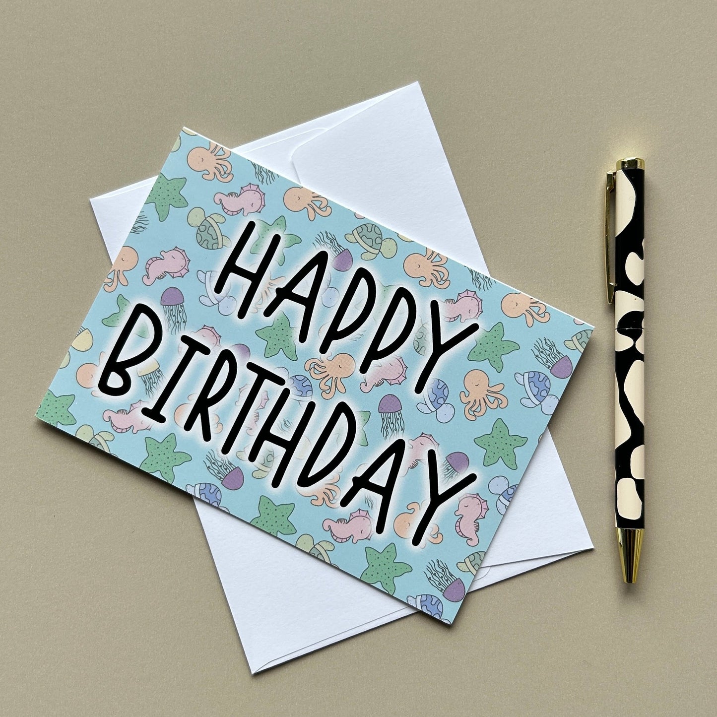A square birthday card with a pastel under the sea pattern and the words 'HAPPY BIRTHDAY' in black script, placed on top of a white envelope. A black-and-white patterned pen with a gold tip rests to the right, all on a warm beige background.