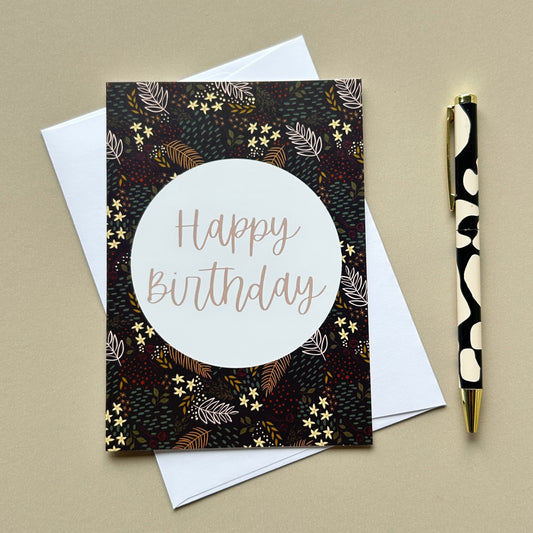 A square birthday card with a dark floral pattern and a large white circle reading 'Happy Birthday' in rose-gold lettering, styled on top of white envelopes with a black-and-white patterned pen on a beige background.