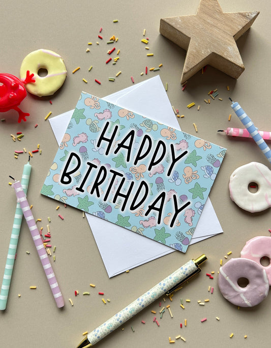 A square birthday card with a pastel under the sea pattern and the words 'HAPPY BIRTHDAY' in black script, placed on top of a white envelope. The card is styled in a flat-lay arrangement with pastel striped straws, a wooden star, donut-shaped cookies, sprinkles, and a pen on a beige background.