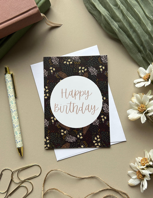 A styled flat-lay of a square birthday card with a dark floral border and a white circle reading 'Happy Birthday' in rose-gold lettering. The card is placed on white paper with decorative props including a pen, paper clips, twine, flowers, notebooks, and a pleated green fabric on a beige surface.