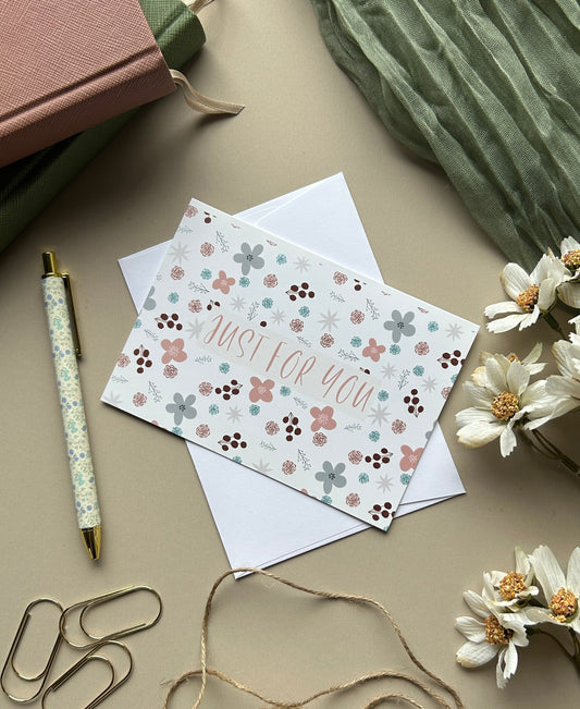 A styled flat-lay featuring a floral 'Just for You' greeting card with a white envelope, surrounded by a pink notebook, green fabric, a patterned pen, paperclips, twine, and white flowers on a neutral beige background.