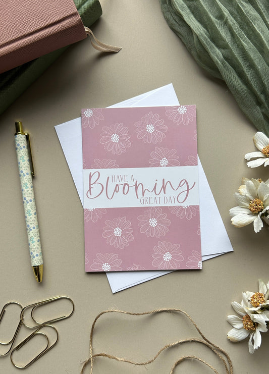 A pink greeting card with white floral illustrations and the words 'Have a Blooming Great Day!' in script, styled on a beige surface with a white envelope, pen, paper clips, twine, flowers, and notebooks.