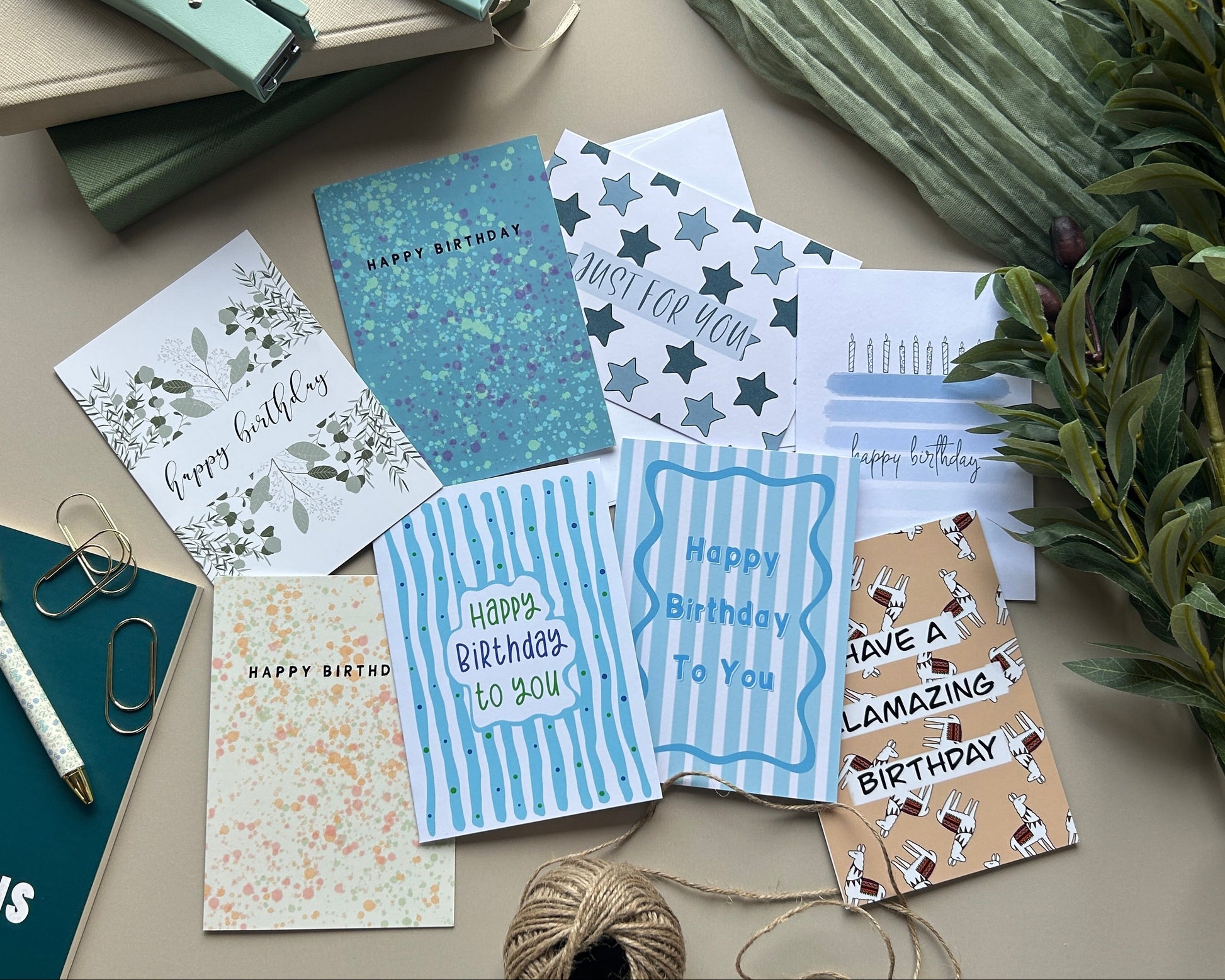 A styled flat-lay of assorted greeting cards and stationery on a neutral beige surface, including blue striped cards, a glittery teal card, a white card with floral writing, and other paper goods alongside notebooks, a pen, paper clips, twine, and greenery.