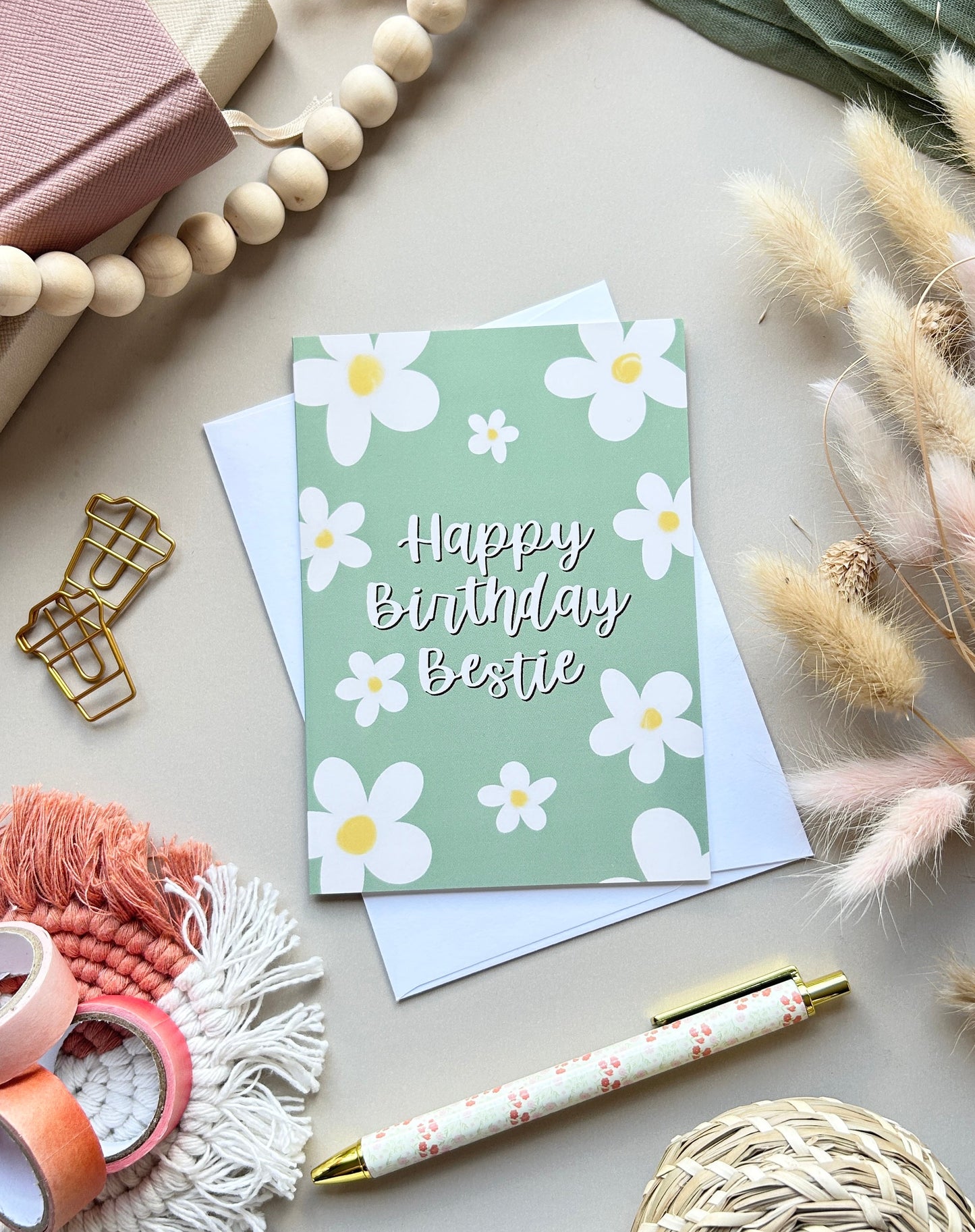 A styled flat-lay featuring a green 'Happy Birthday Bestie' greeting card with white daisy illustrations, accompanied by a white envelope, a floral pen, and various boho decorative props on a neutral background.