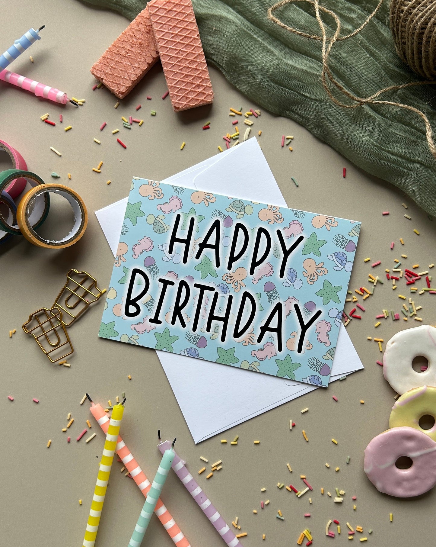 A square birthday card with a pastel under the sea pattern and the words 'HAPPY BIRTHDAY' in black lettering, placed on top of a white envelope. The card is styled in a flat lay with pastel pencils, ribbon, confetti, cookies, and fabric on a neutral surface.