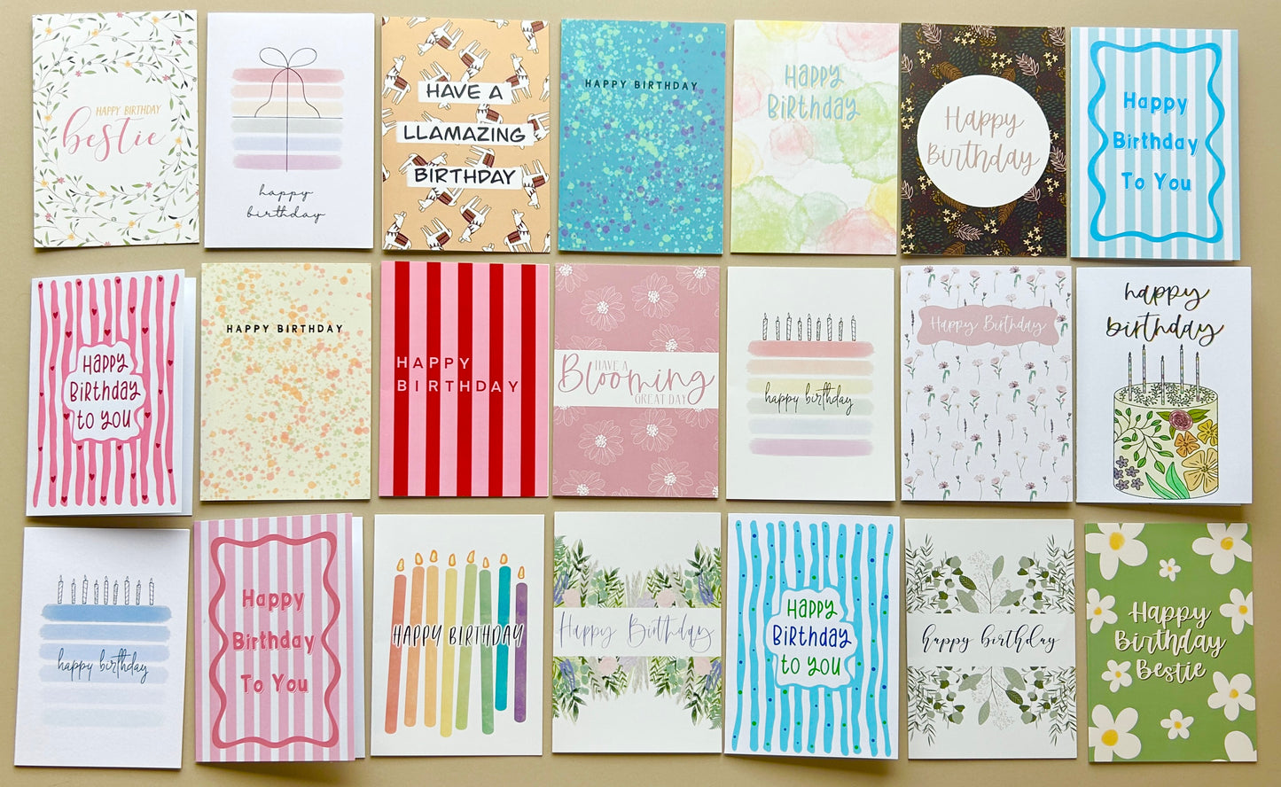 A 3x7 grid of 21 different birthday greeting cards arranged on a light beige surface. The cards feature various colourful designs, including stripes, watercolour textures, floral prints, and birthday cake illustrations, with text such as 'Happy Birthday', 'Have a LLAMazing Birthday', and 'Happy Birthday To You'.