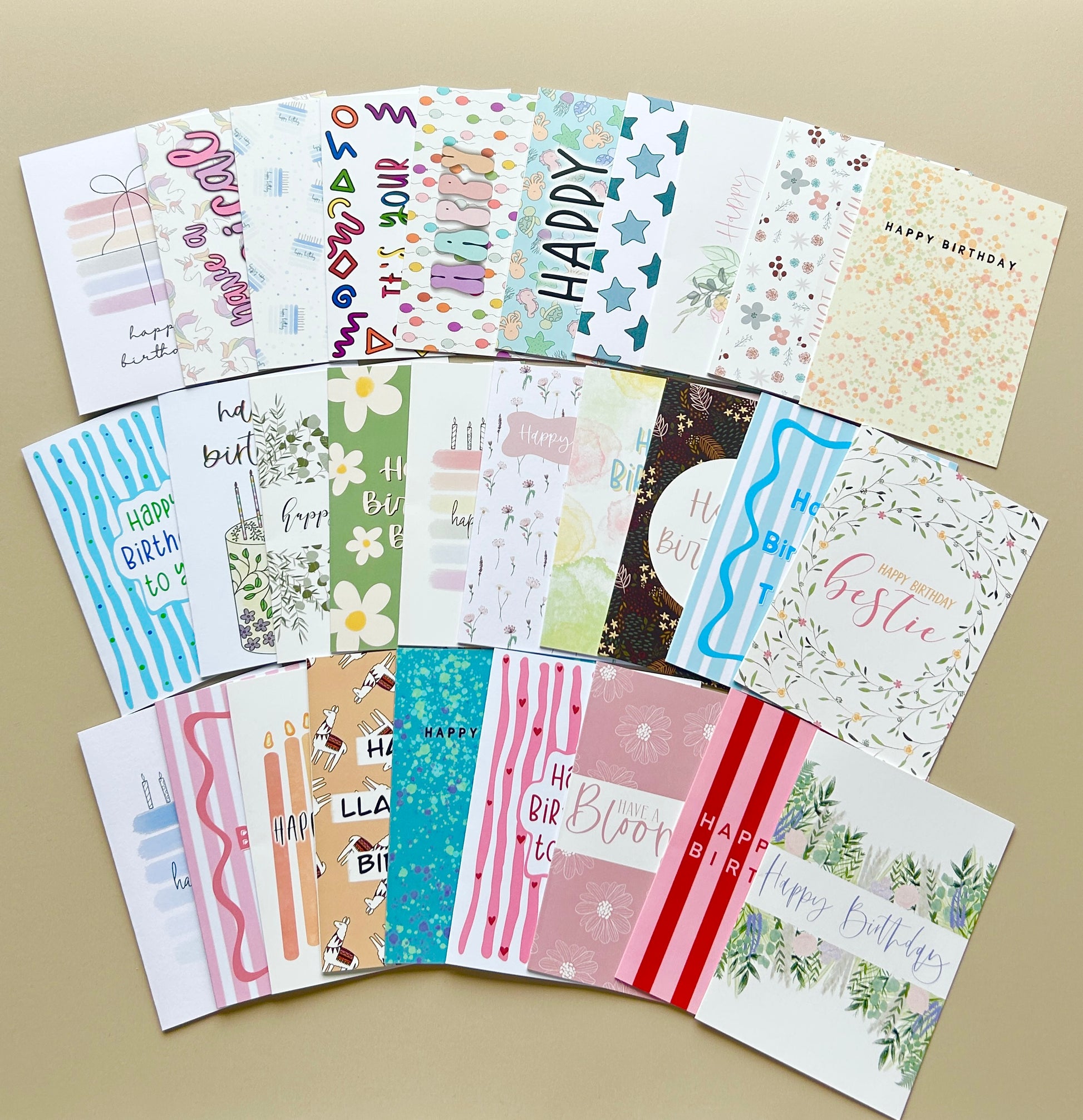 A set of colorful birthday greeting cards arranged in a fan shape on a beige background. The cards feature various patterns including stripes, florals, stars, and illustrations, with 'Happy Birthday' and 'HBD' written on several designs.