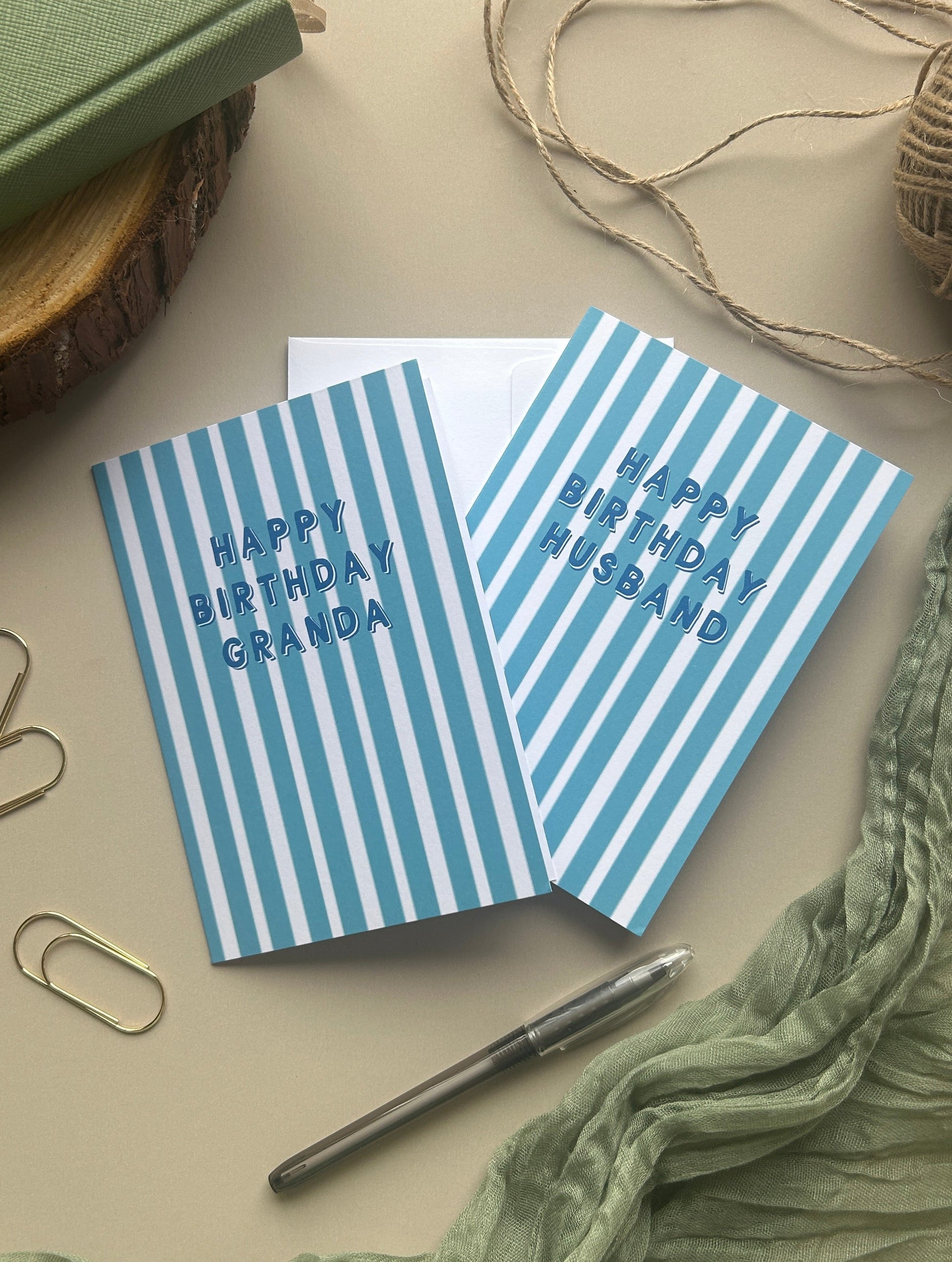 Two striped birthday greeting cards with blue and white vertical stripes, one reading 'HAPPY BIRTHDAY GRANDPA' and the other 'HAPPY BIRTHDAY HUSBAND', styled on a beige surface with a green notebook, twine, paper clips, a pen, and a crinkled green fabric.