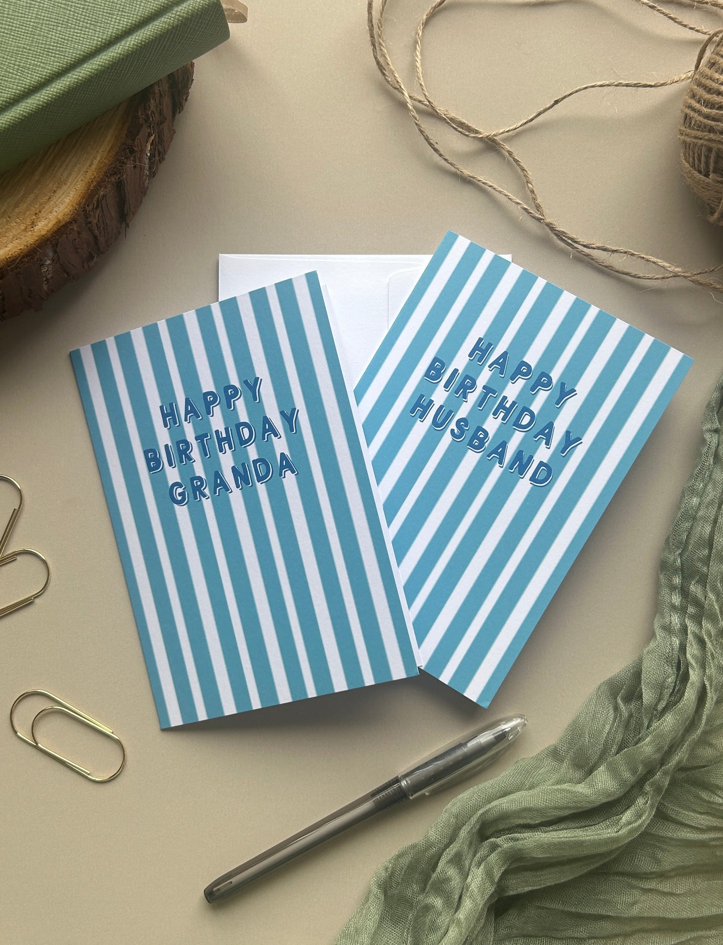 Two striped birthday greeting cards with blue and white vertical stripes, one reading 'HAPPY BIRTHDAY GRANDPA' and the other 'HAPPY BIRTHDAY HUSBAND', styled on a beige surface with a green notebook, twine, paper clips, a pen, and a crinkled green fabric.