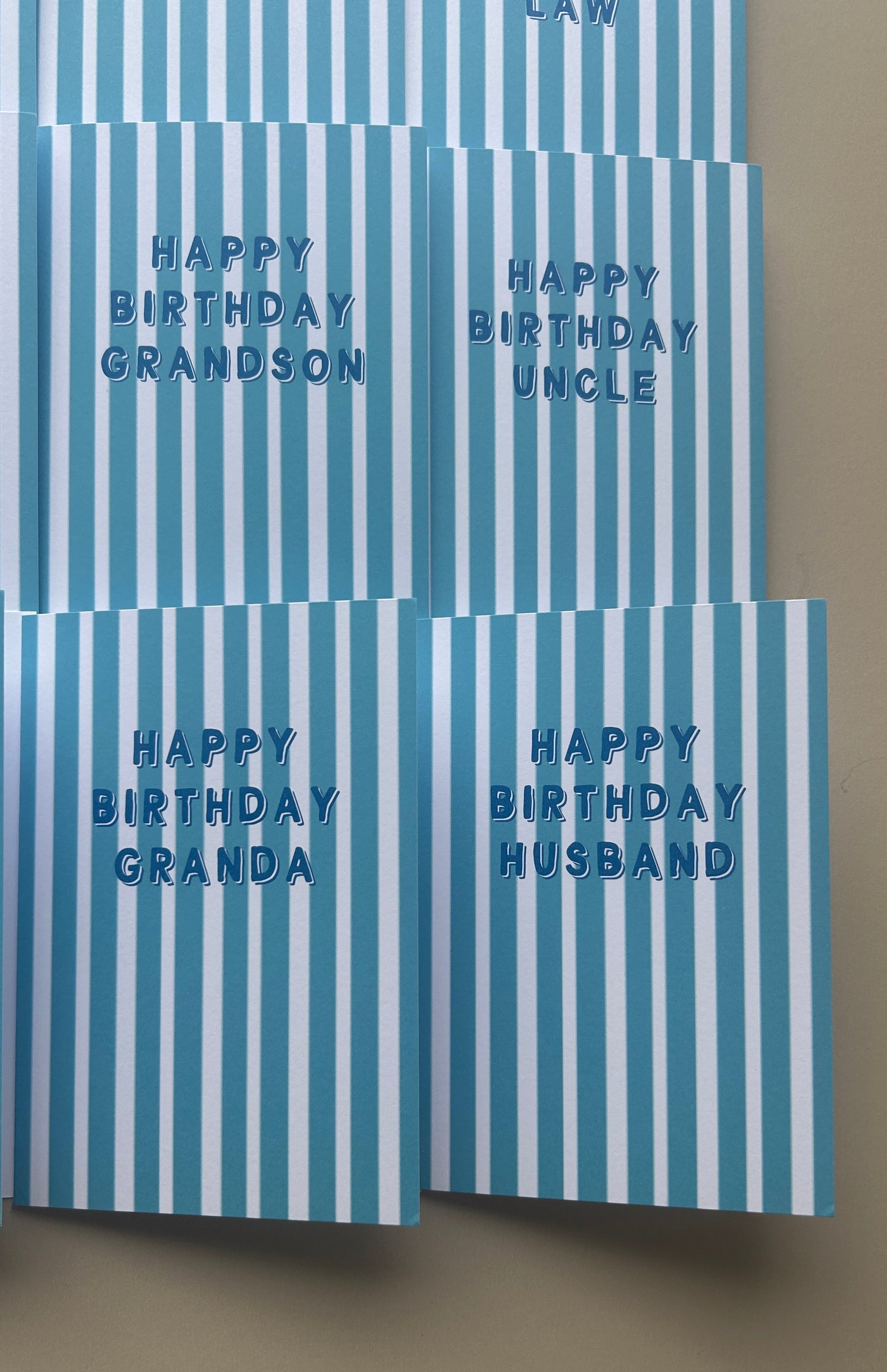 Four striped greeting cards arranged in a grid on a beige surface. The cards have vertical light blue and white stripes and are personalized with blue text reading 'HAPPY BIRTHDAY GRANDSON,' 'HAPPY BIRTHDAY UNCLE,' 'HAPPY BIRTHDAY GRANDA,' and 'HAPPY BIRTHDAY HUSBAND.