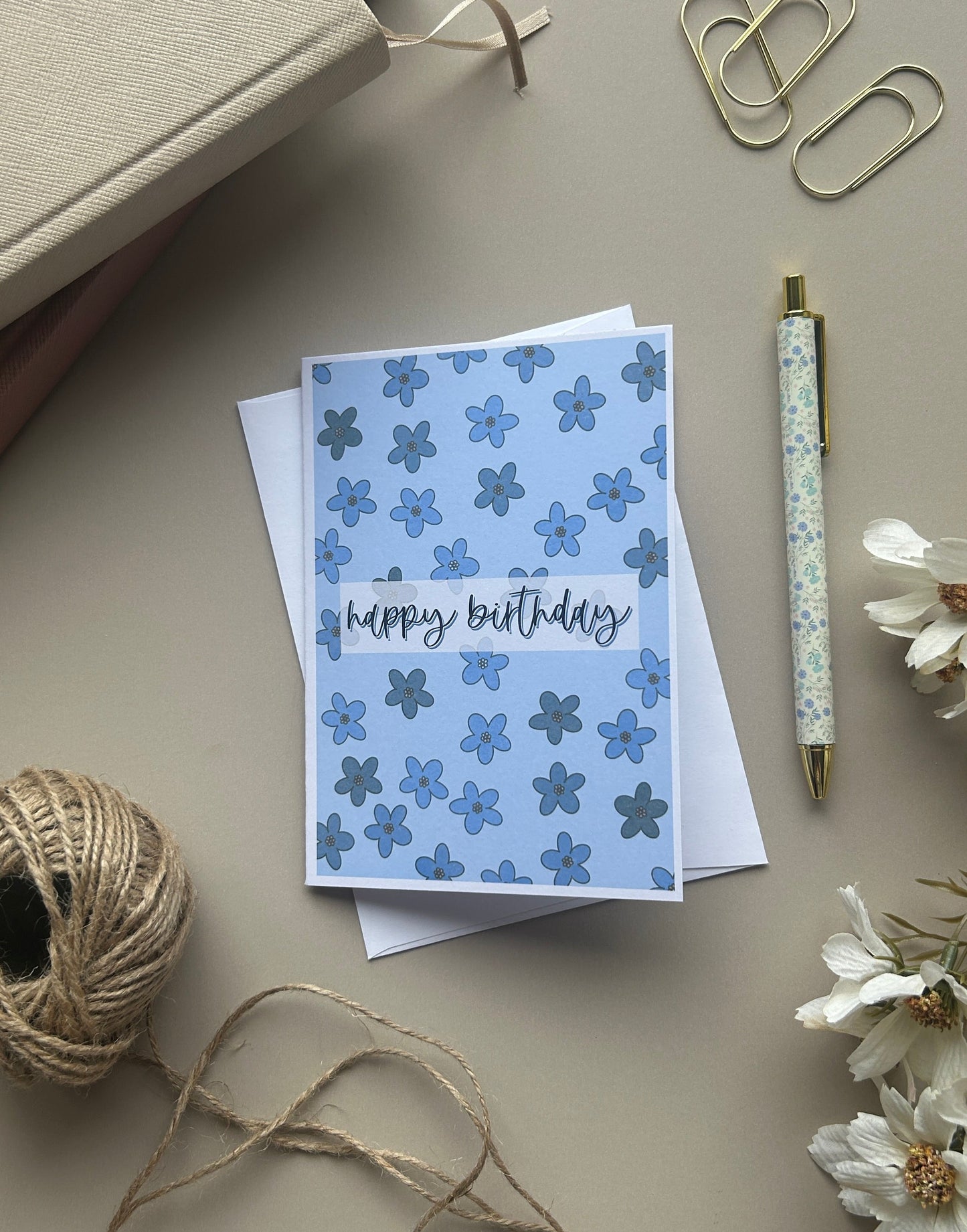 A blue floral 'happy birthday' greeting card with white and blue flowers, styled on a beige desk with a pen, paper clips, twine, and dried flowers.