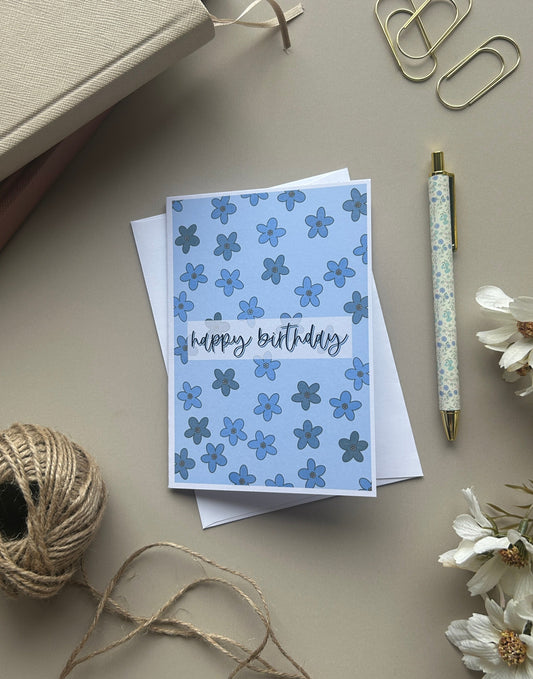 A blue floral 'happy birthday' greeting card with white and blue flowers, styled on a beige desk with a pen, paper clips, twine, and dried flowers.