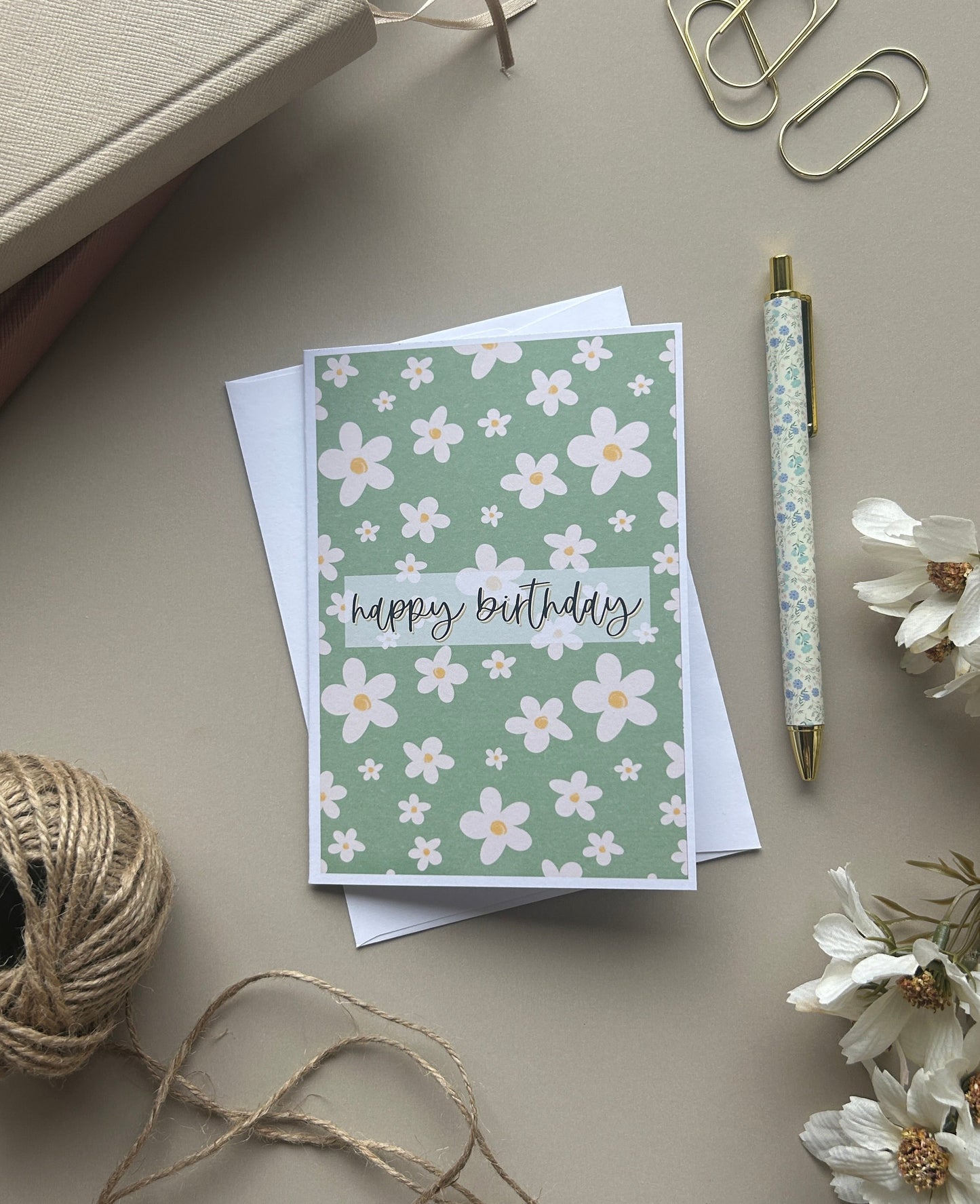 A floral greeting card with 'happy birthday' written in script, styled in a flat lay with a pen, paper clips, twine, dried flowers, and a notebook on a neutral beige background.