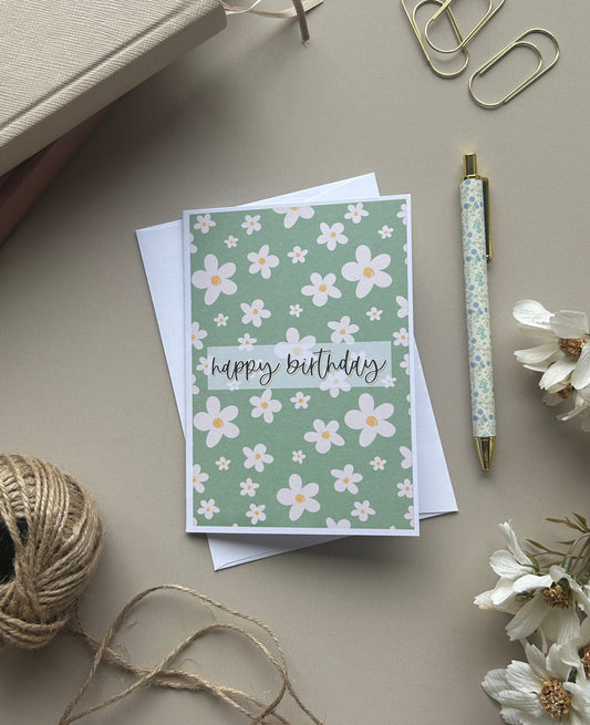 A floral greeting card with 'happy birthday' written in script, styled in a flat lay with a pen, paper clips, twine, dried flowers, and a notebook on a neutral beige background.