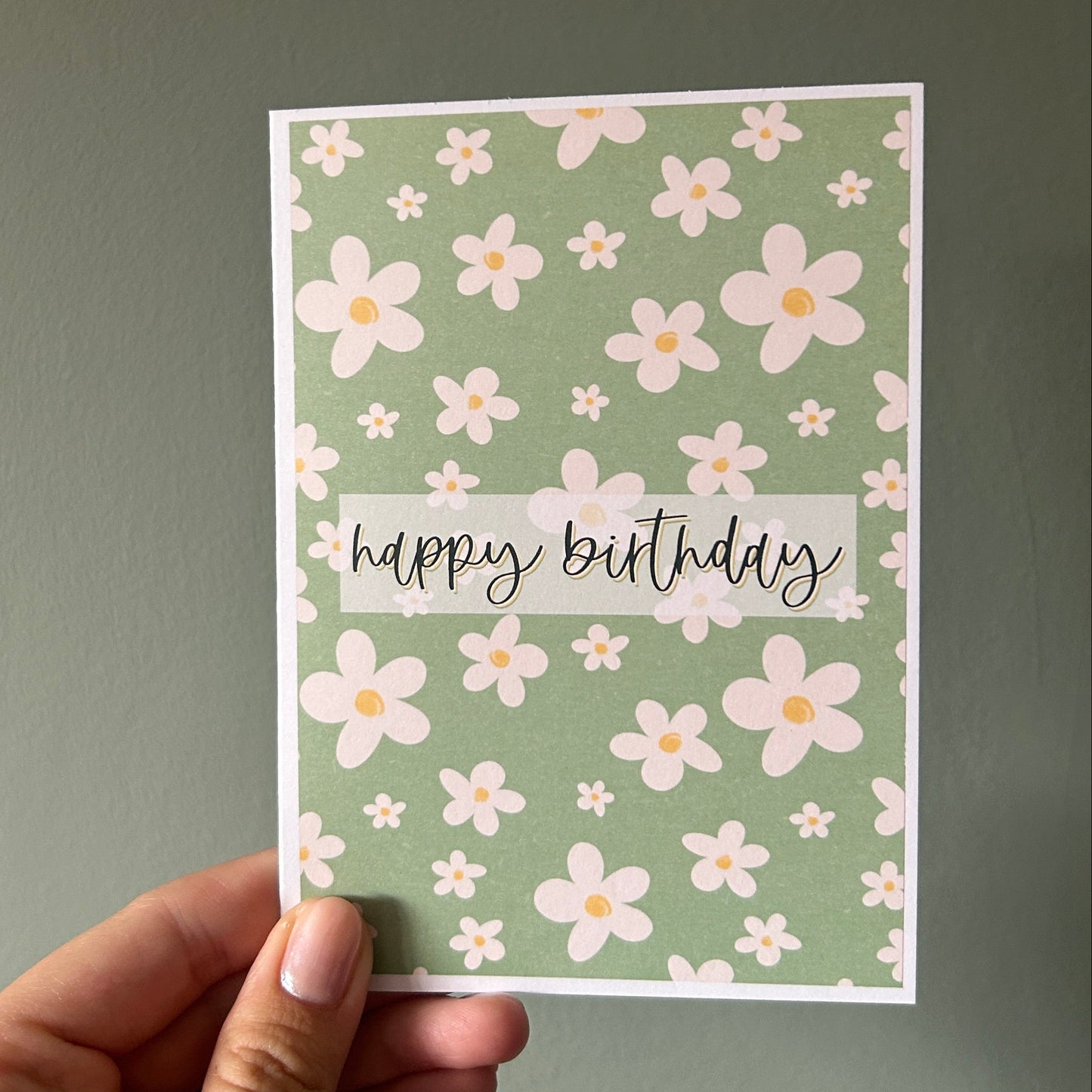 A hand holding a handmade square greeting card with a sage green background, white and pink daisy pattern, and a white banner reading 'happy birthday' in black script, set against a solid dark gray wall.
