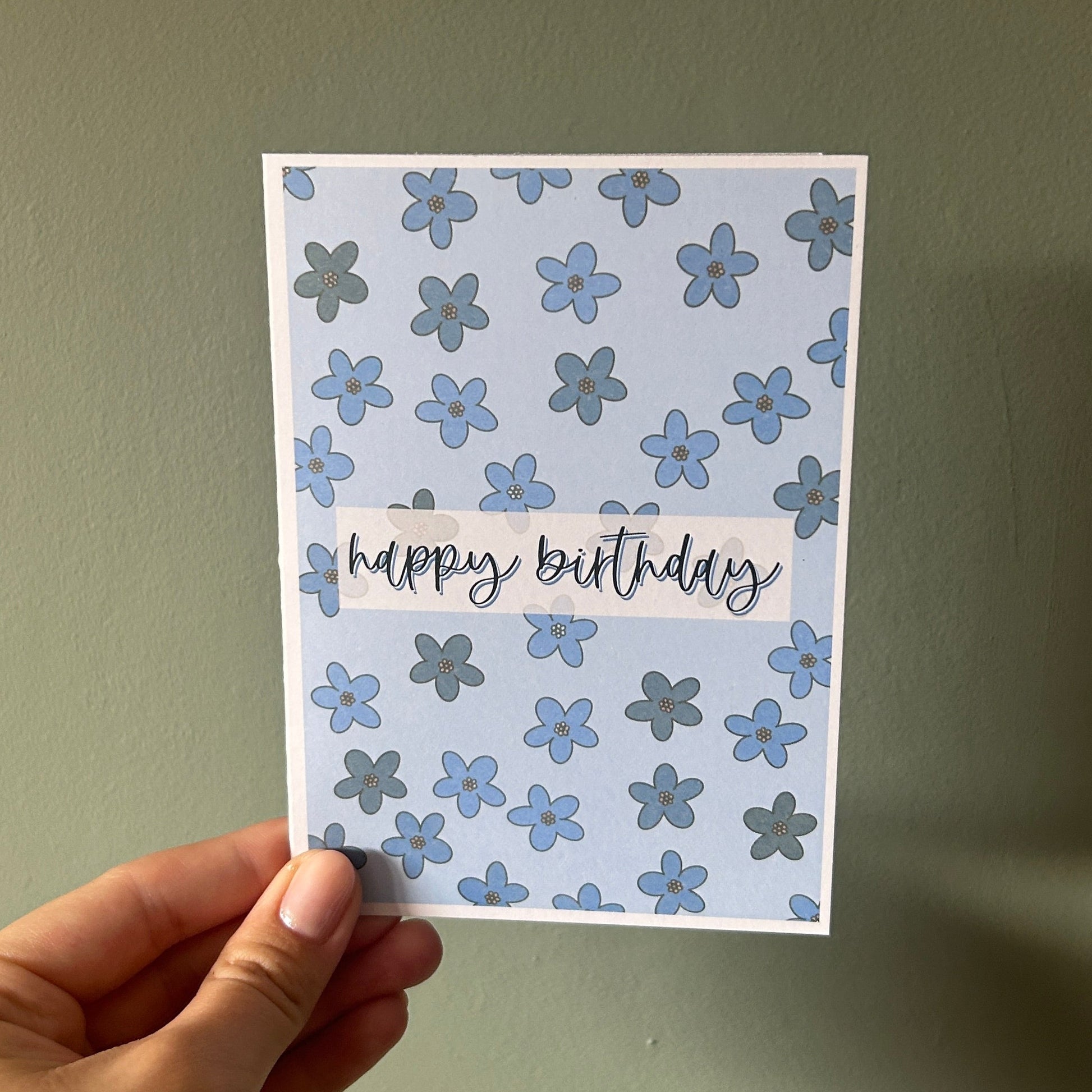 A hand holding a square greeting card with a light blue and white floral pattern and the words 'happy birthday' in black script, against a muted green wall.