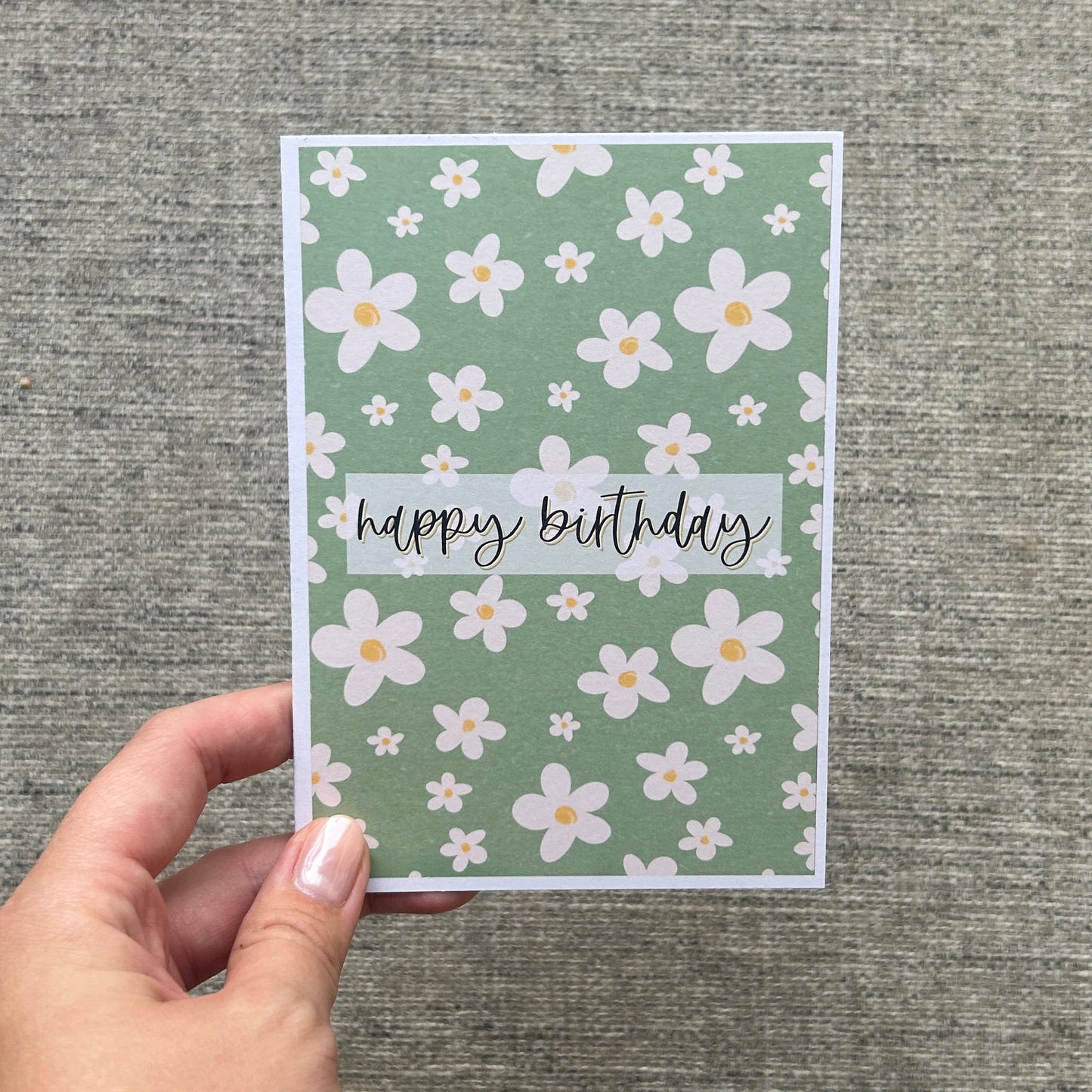 A hand with white nail polish and gold bracelets holds a square greeting card with a sage green floral pattern and the words 'happy birthday' on a textured grey background.