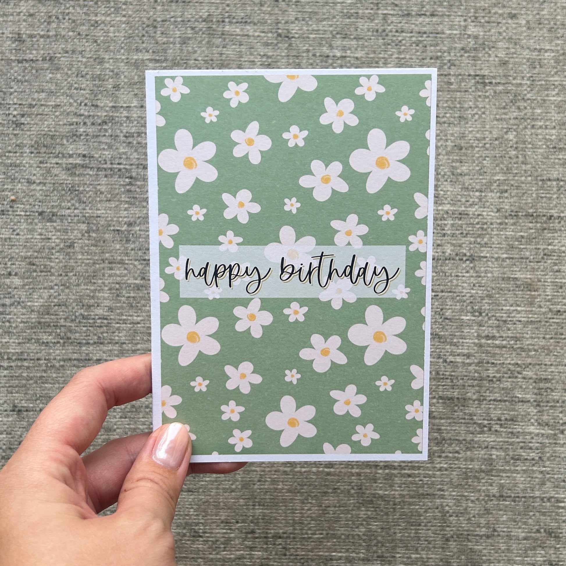 A hand with white nail polish and gold bracelets holds a square greeting card with a sage green floral pattern and the words 'happy birthday' on a textured grey background.