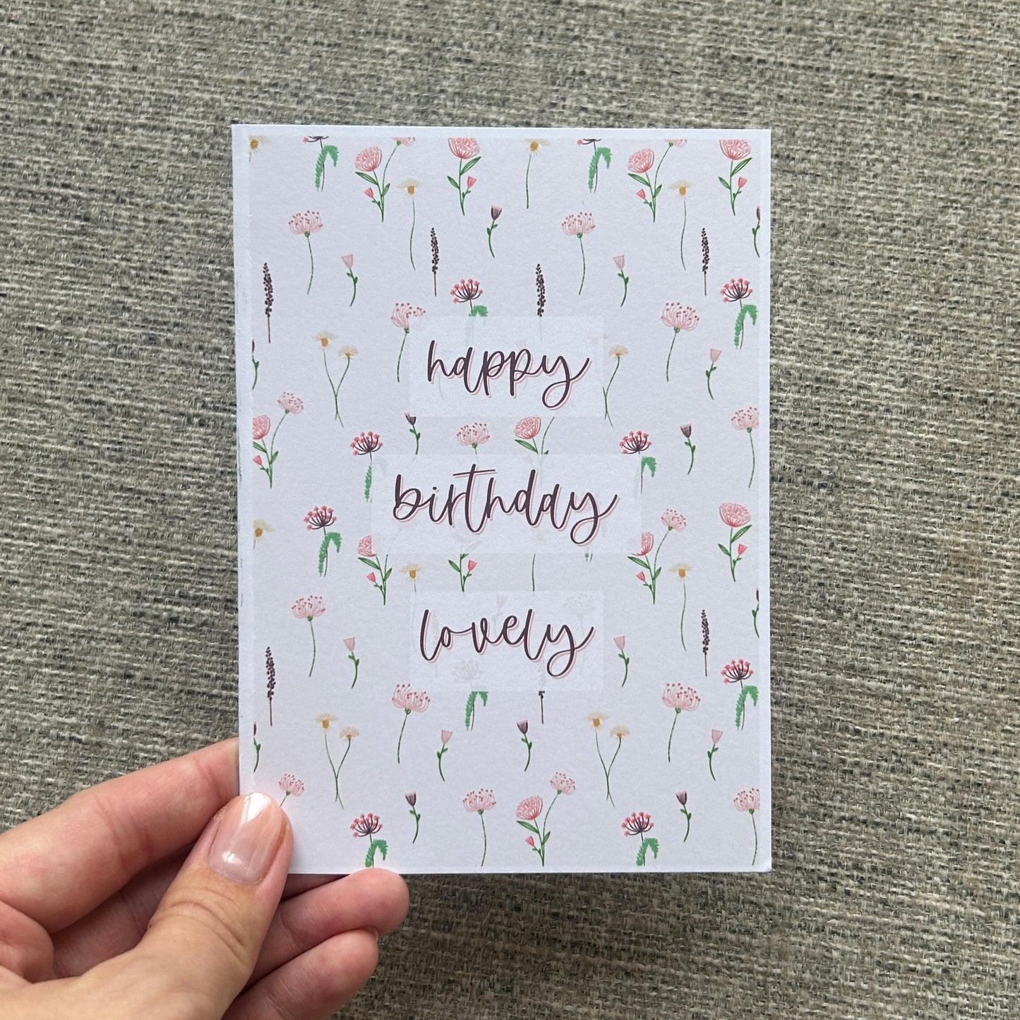 A hand holding a square greeting card with a white background, decorated with a delicate pink and green floral pattern and the words 'happy birthday lovely' in a cursive font, set against a textured gray fabric background.